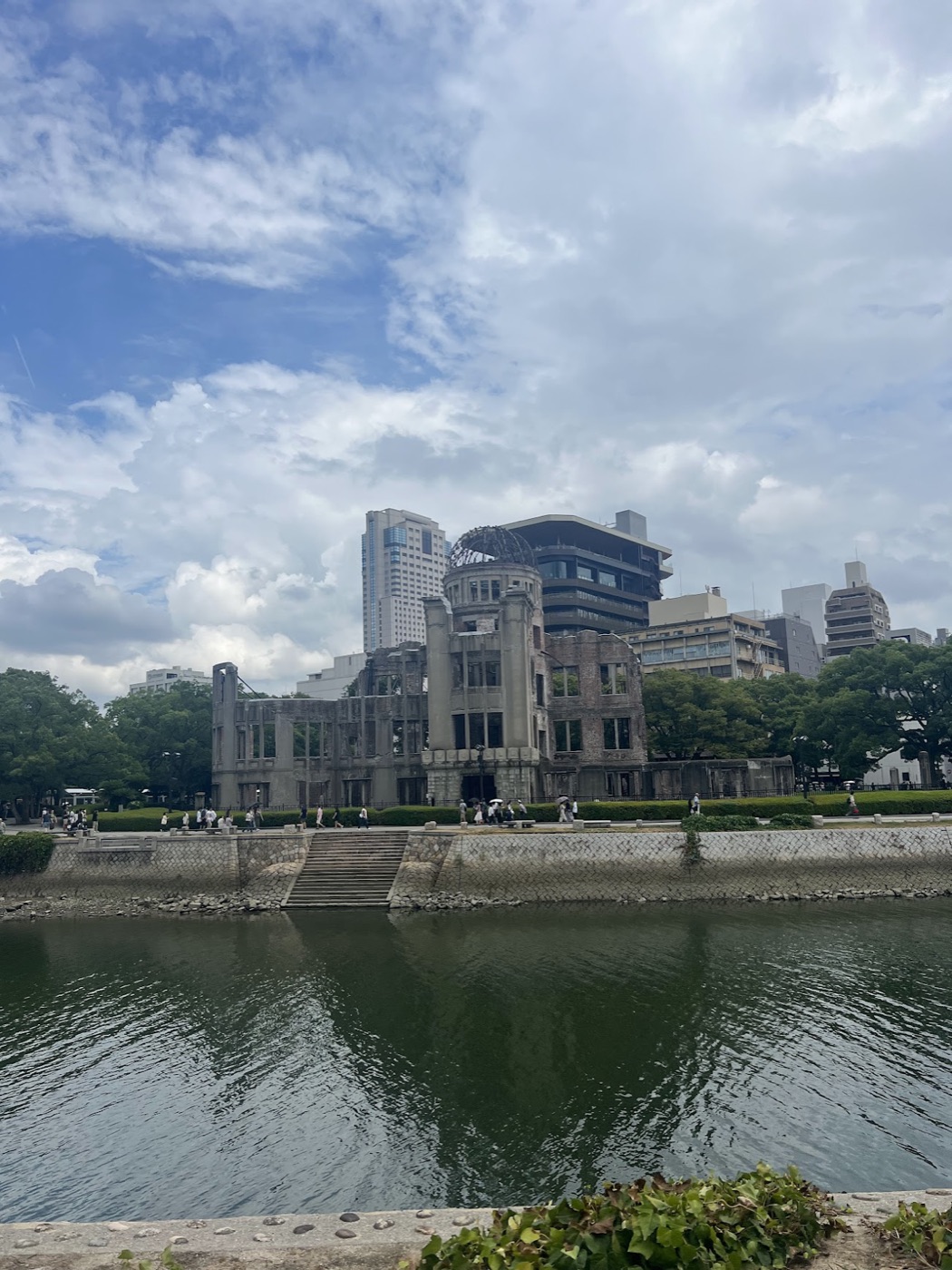The ruined Atomic Bomb Dome in Hiroshima stands across the river under a bright, cloud-streaked sky.