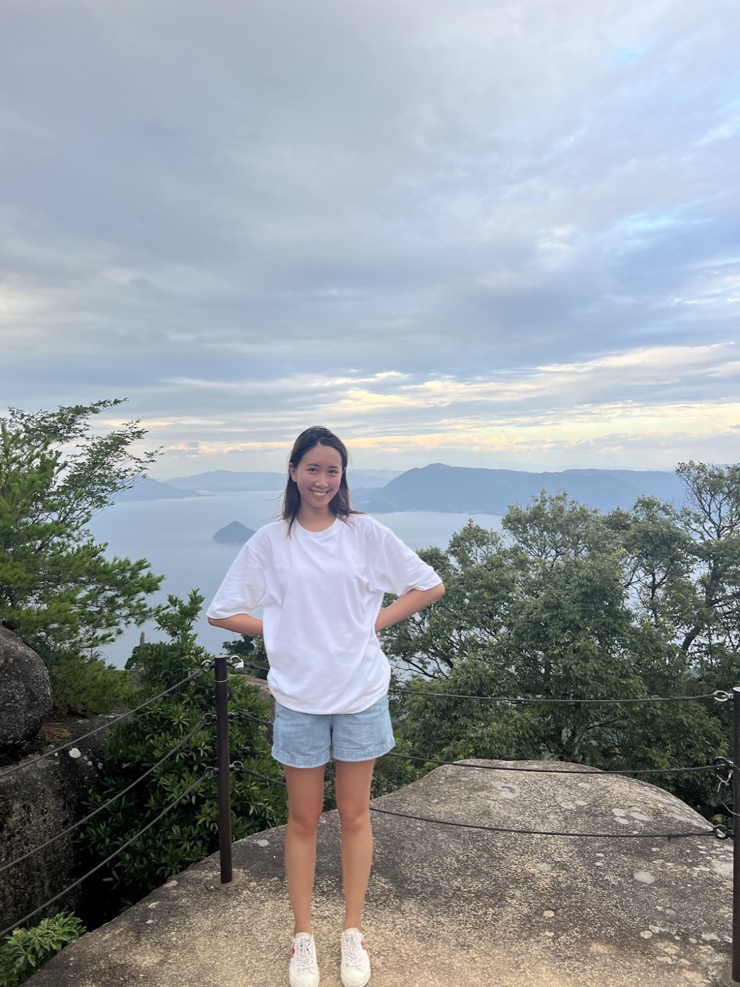 A woman in a white tee and denim shorts stands at a roped stone overlook with forested islands and a hazy sea behind her.