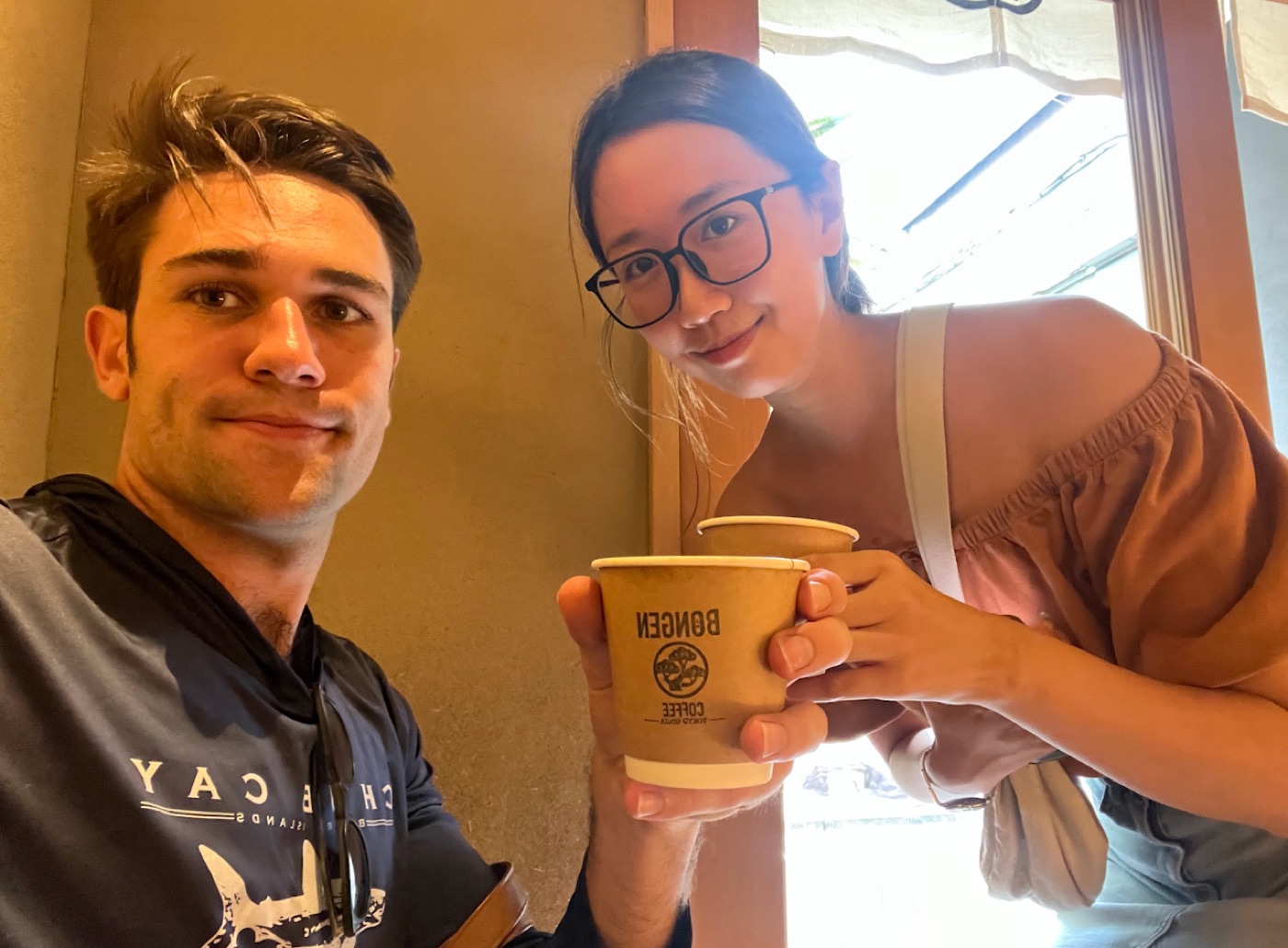 Selfie of Noel and a woman in glasses raising paper coffee cups in a sunlit doorway.