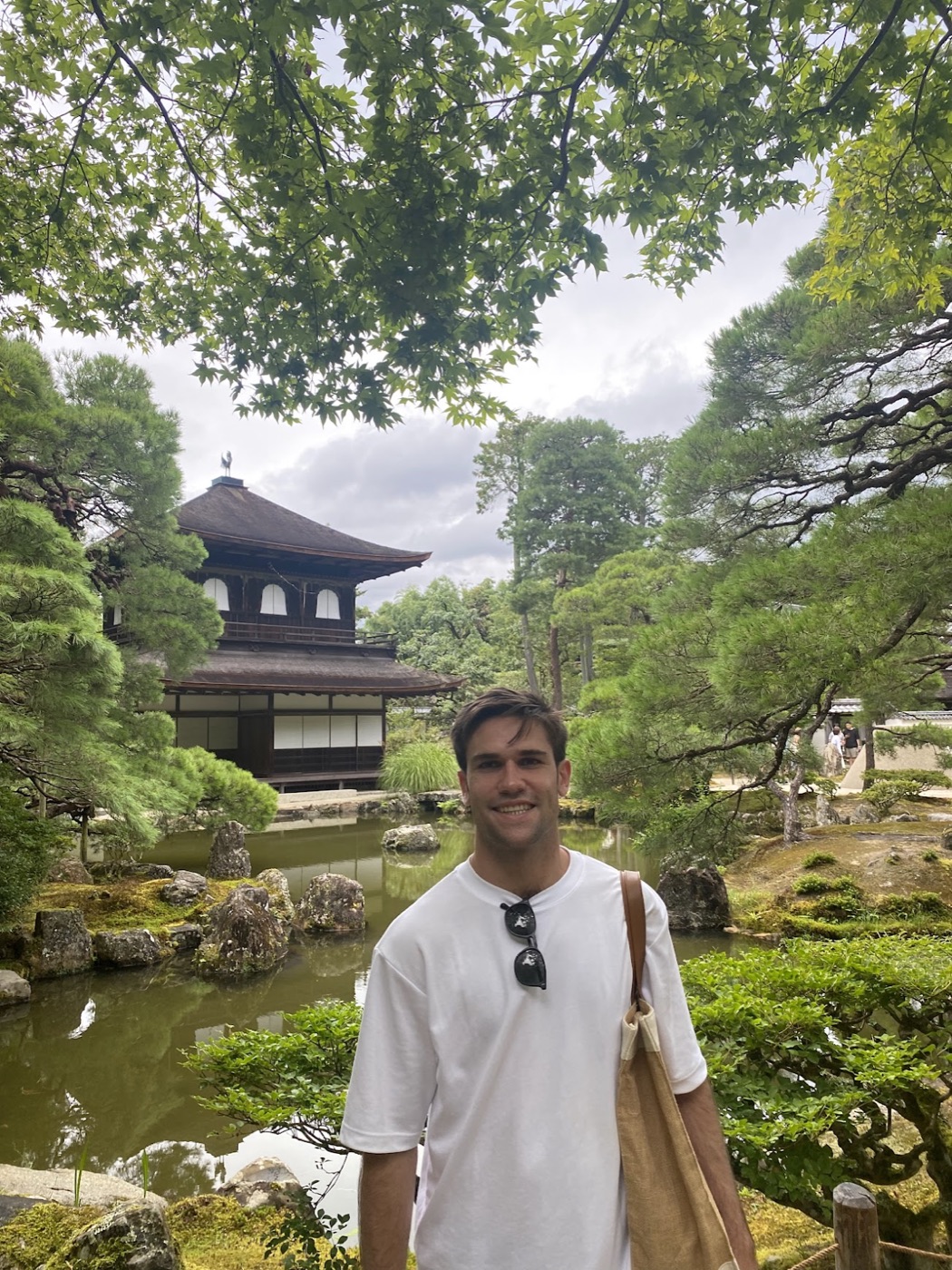 Noel in a white tee smiles in front of a pond and a two-story wooden temple pavilion ringed by pines.