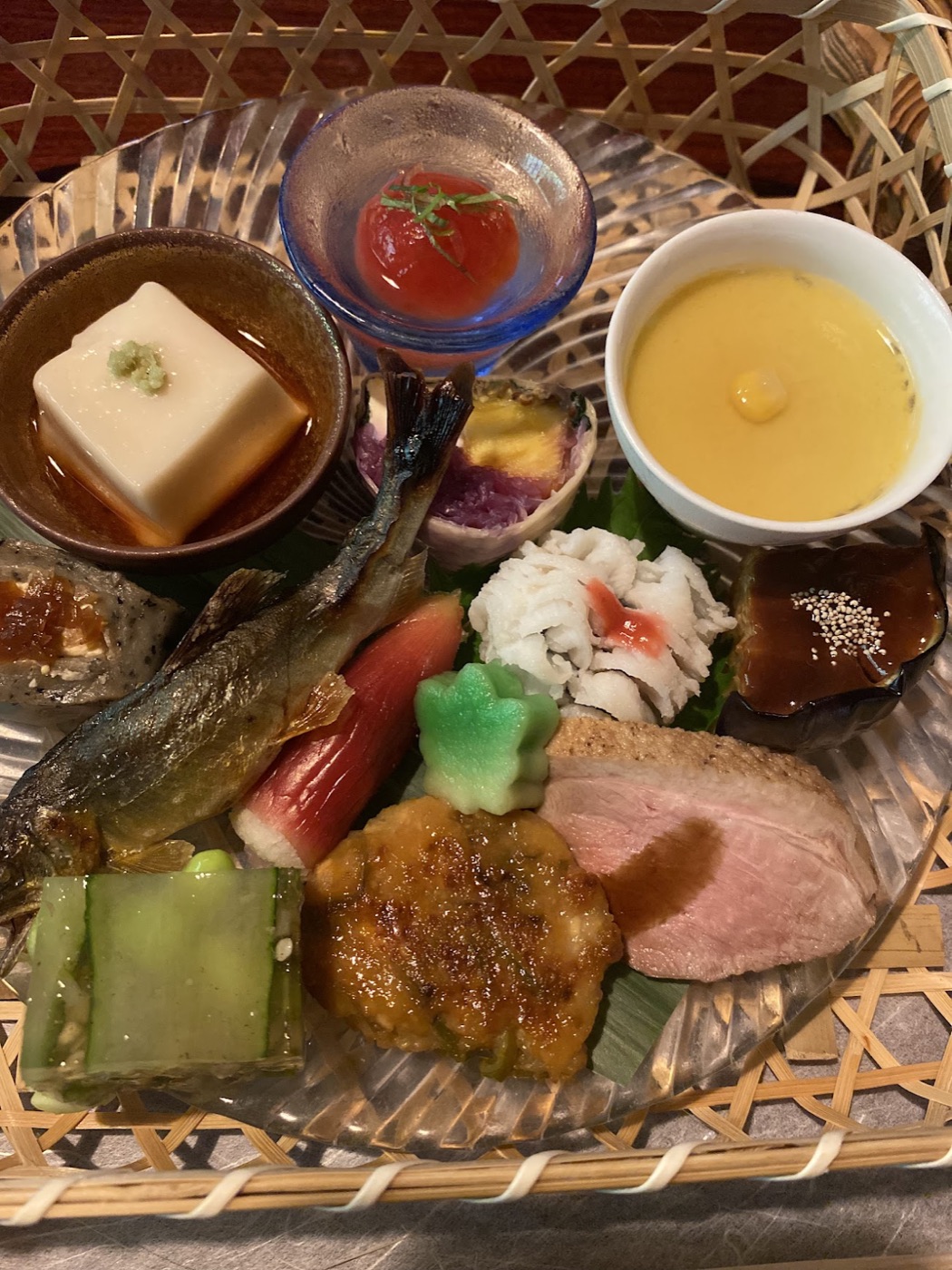A woven basket platter of kaiseki bites including a whole grilled fish, chilled tofu, sliced duck, and a cup of corn soup.