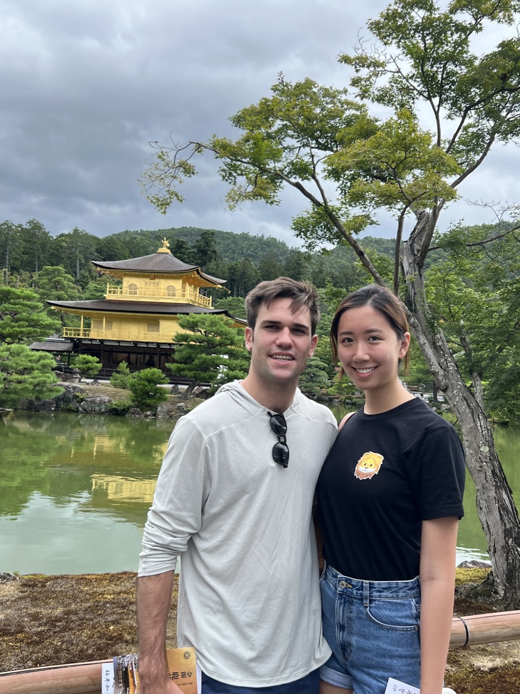 Noel and a woman in a black lion-print tee stand arm in arm before the gold-leafed Kinkaku-ji and its reflecting pond.