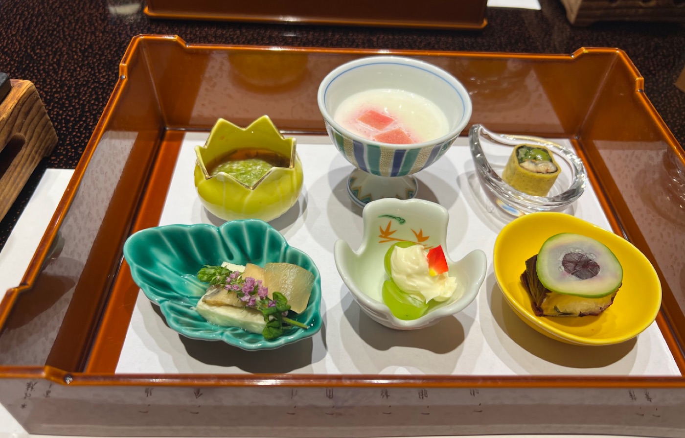 Six small kaiseki starters in mismatched colorful dishes arranged on a lacquered tray.