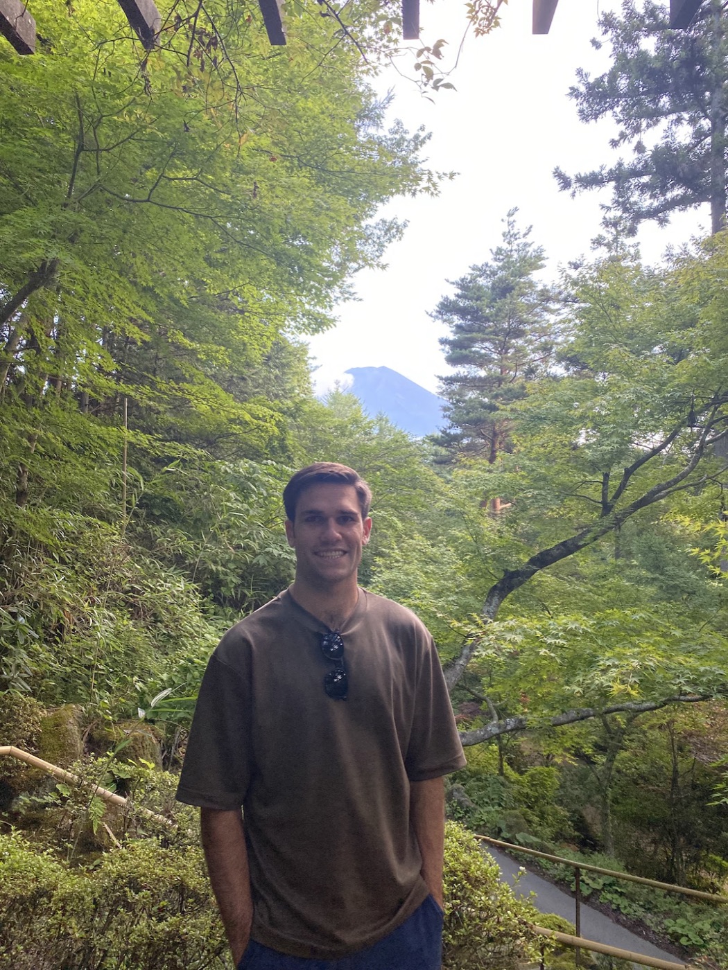 Noel in a brown tee stands on a forest path with Mount Fuji's peak just visible through the trees behind him.