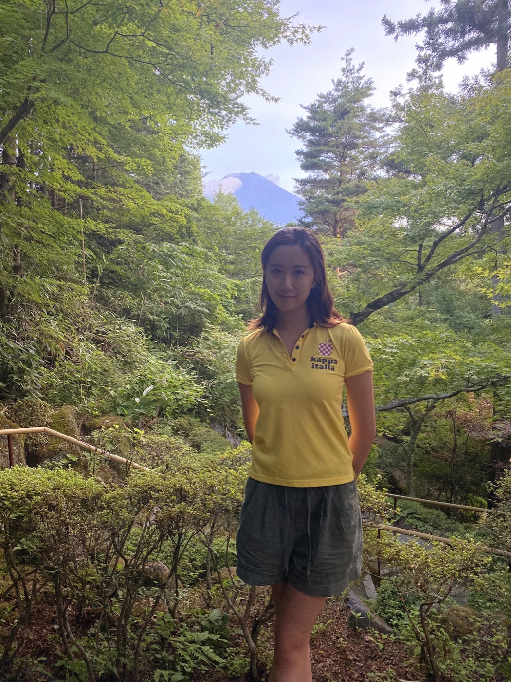 A woman in a yellow polo stands on a wooded trail with Mount Fuji faint in the distance behind her.
