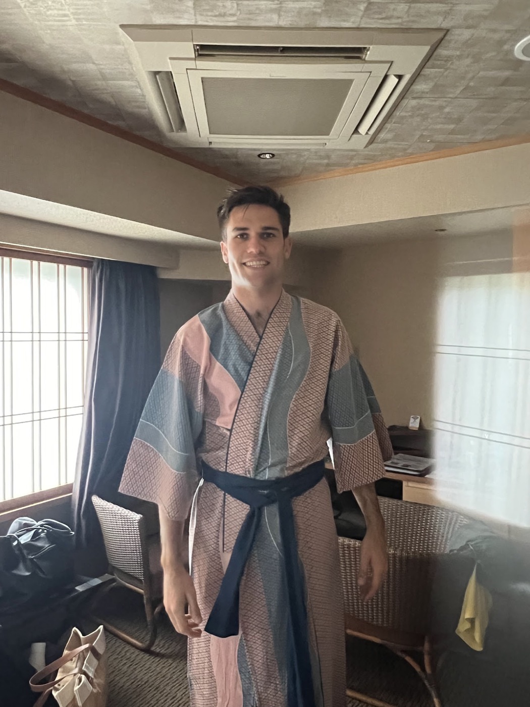 Noel grins in a hotel room wearing a pink-and-gray patterned yukata tied with a navy sash.