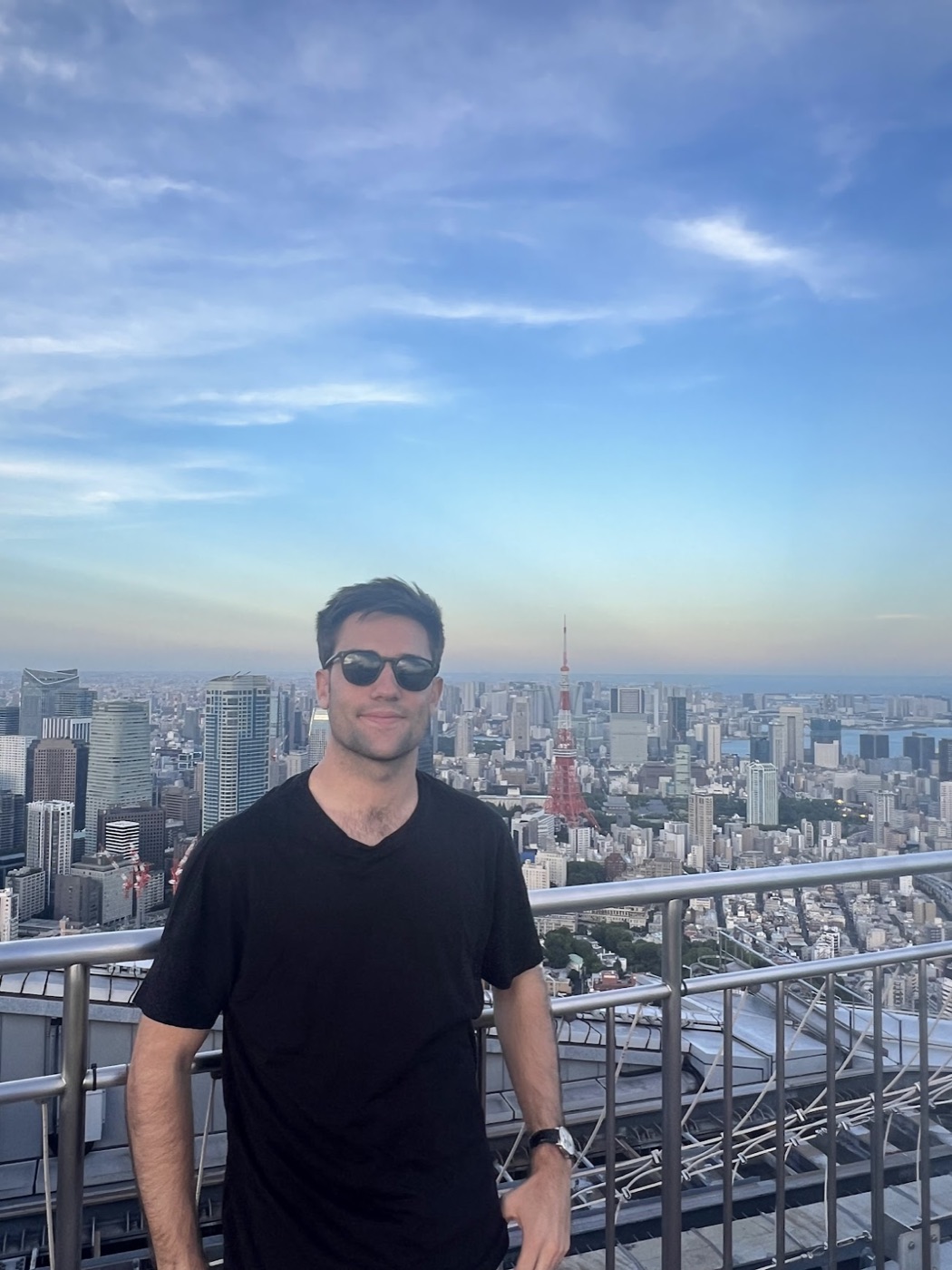 Noel in sunglasses stands on an open-air observation deck with Tokyo Tower and the city sprawling behind him.