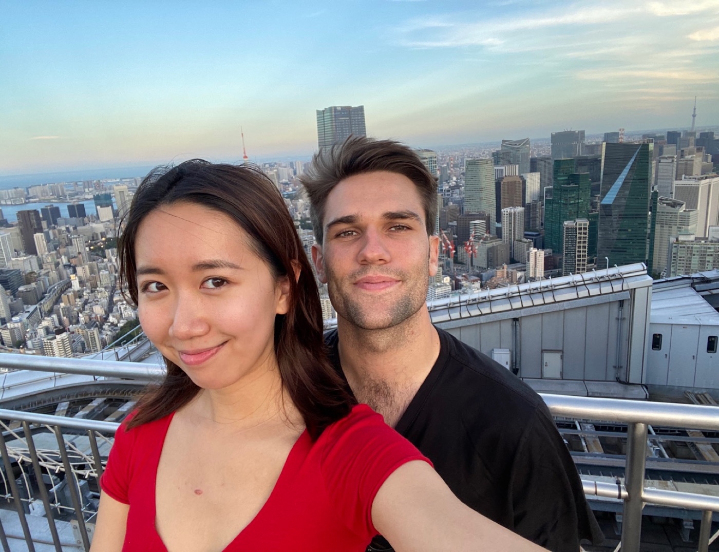 Rooftop selfie of a woman in red and Noel with Tokyo's skyline stretching to the bay behind them.
