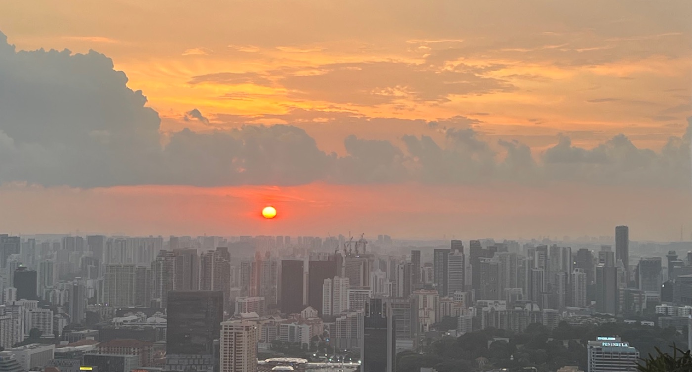 A red sun sinks into orange haze over a dense high-rise skyline at dusk.