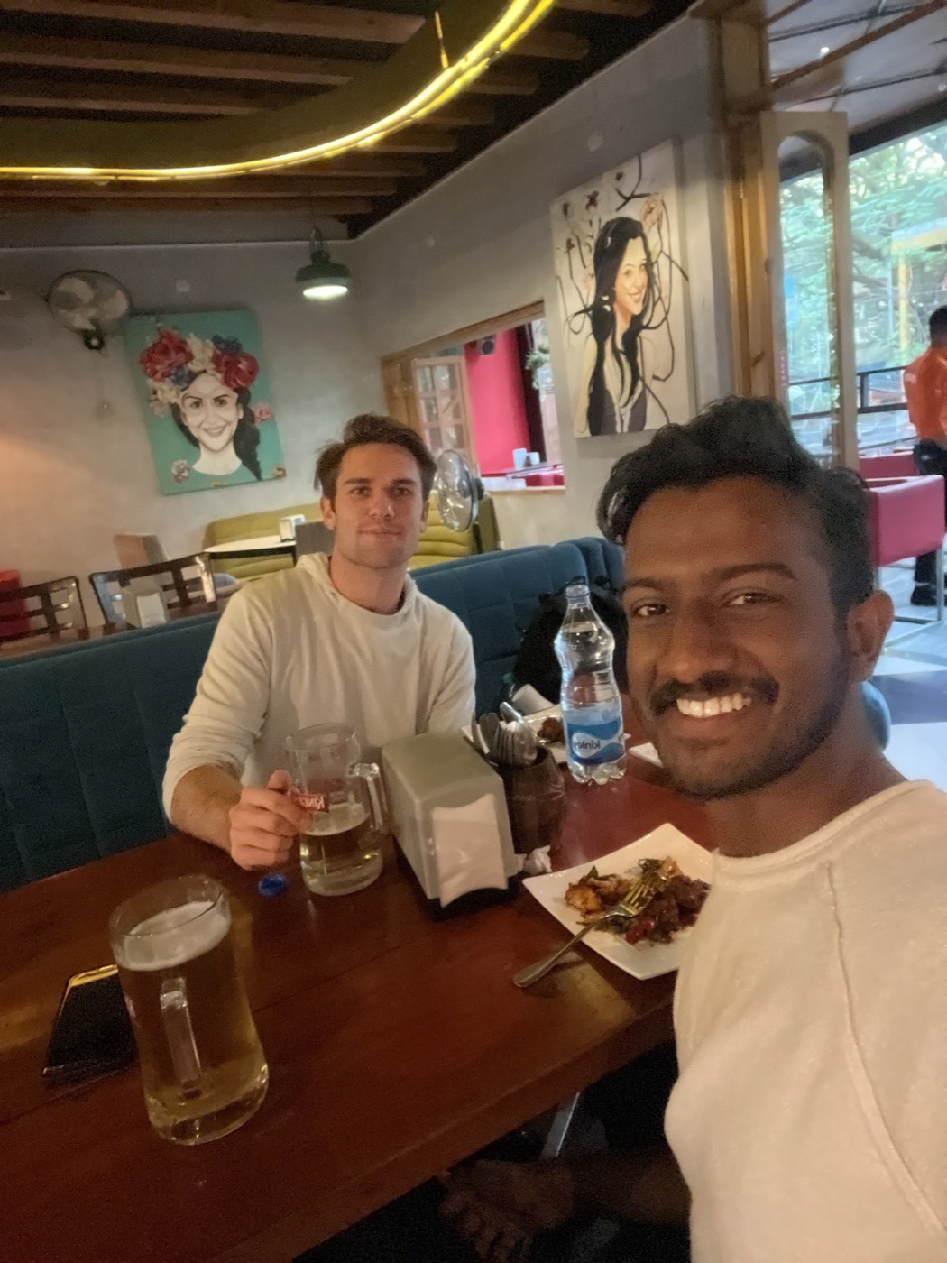 Noel and a mustachioed friend share curry and tall mugs of beer in a colorful cafe hung with portrait paintings.