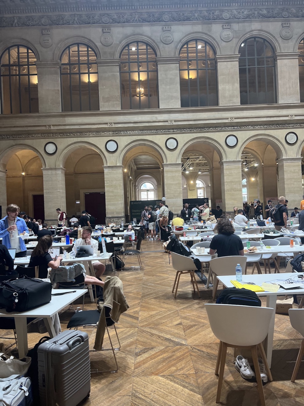 Laptops, luggage, and hackers fill a grand two-story stone hall with arched windows during a hackathon.