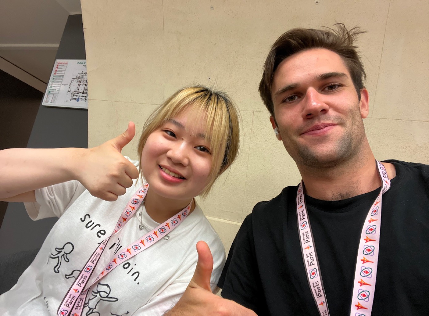 Selfie of Noel and a friend wearing matching pink Paris event lanyards, both giving thumbs up.