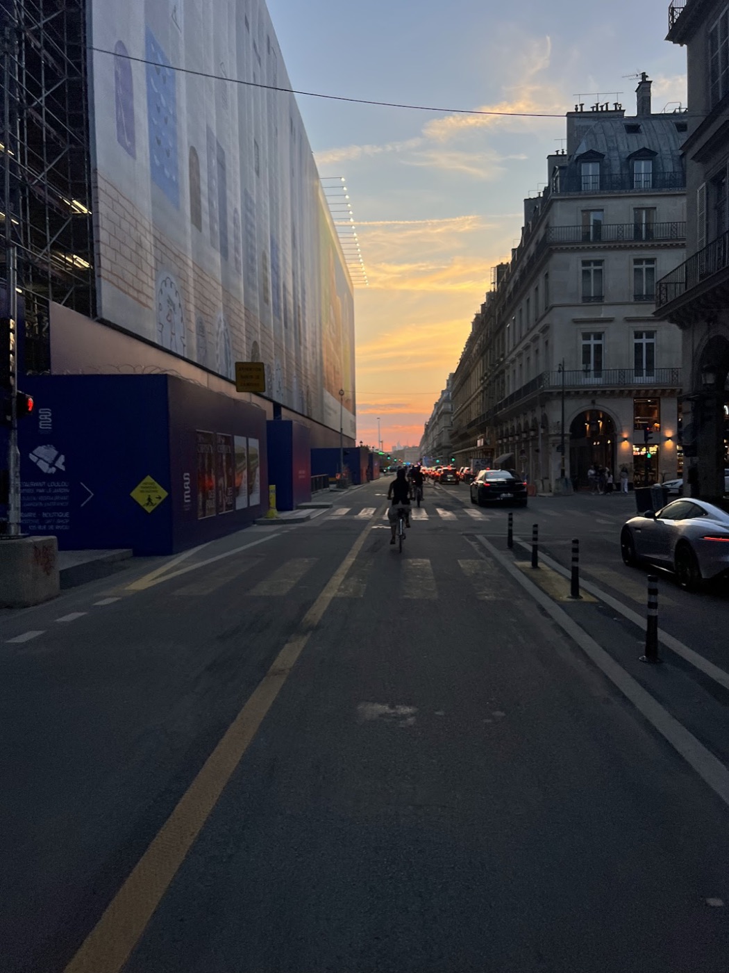 Cyclists ride down a Paris street at sunset between Haussmann facades and a mural-covered construction hoarding.