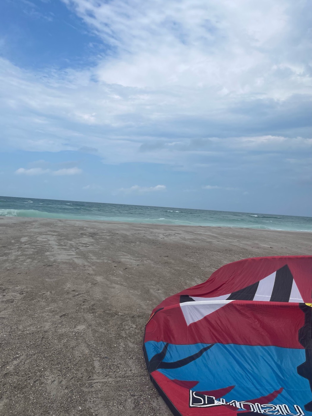A red-and-blue kiteboarding kite lies deflated on brown sand with pale green surf breaking beyond.