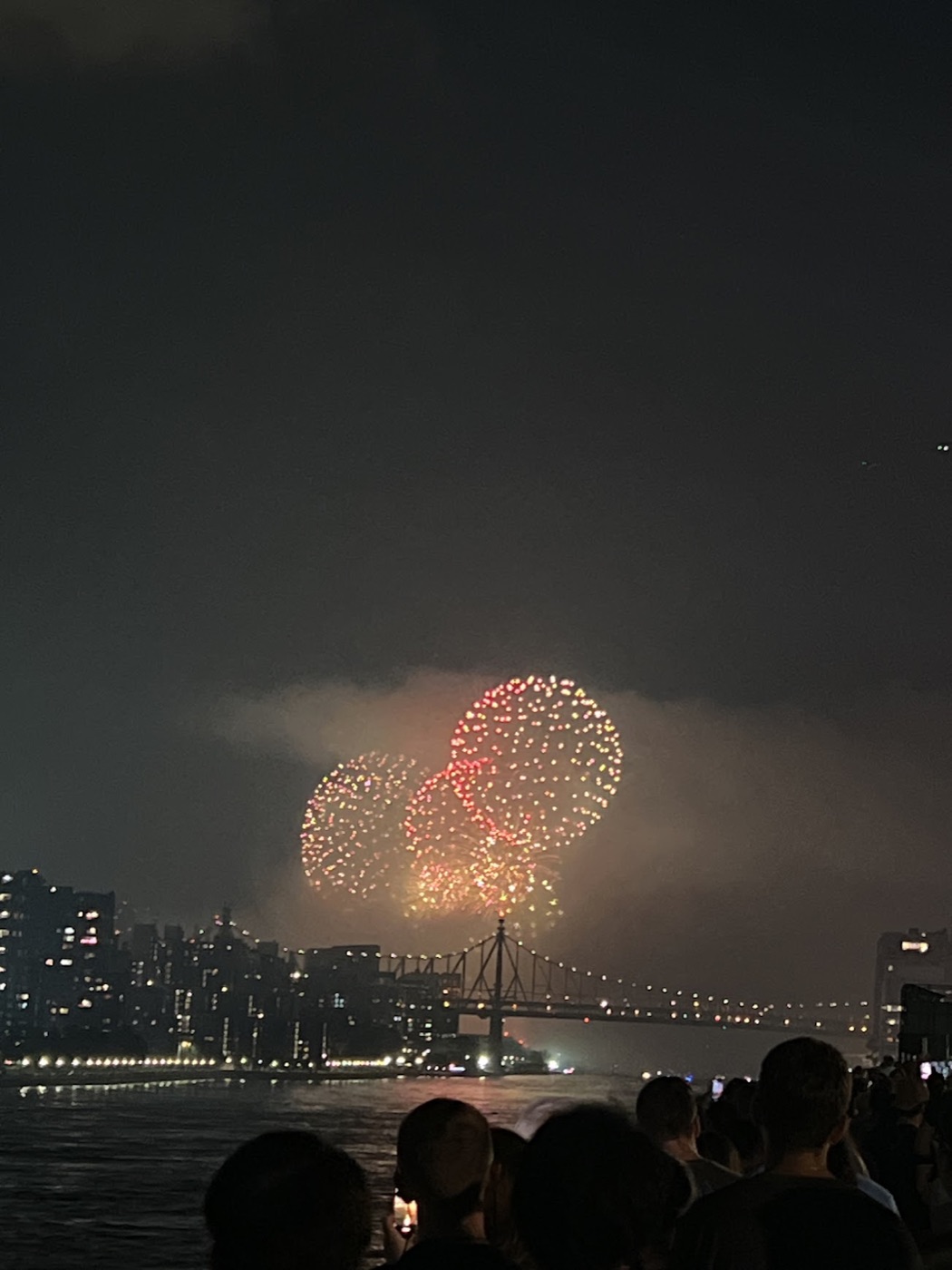 Three fireworks bursts glow through smoke above a lit steel bridge as silhouetted spectators watch from across the river.