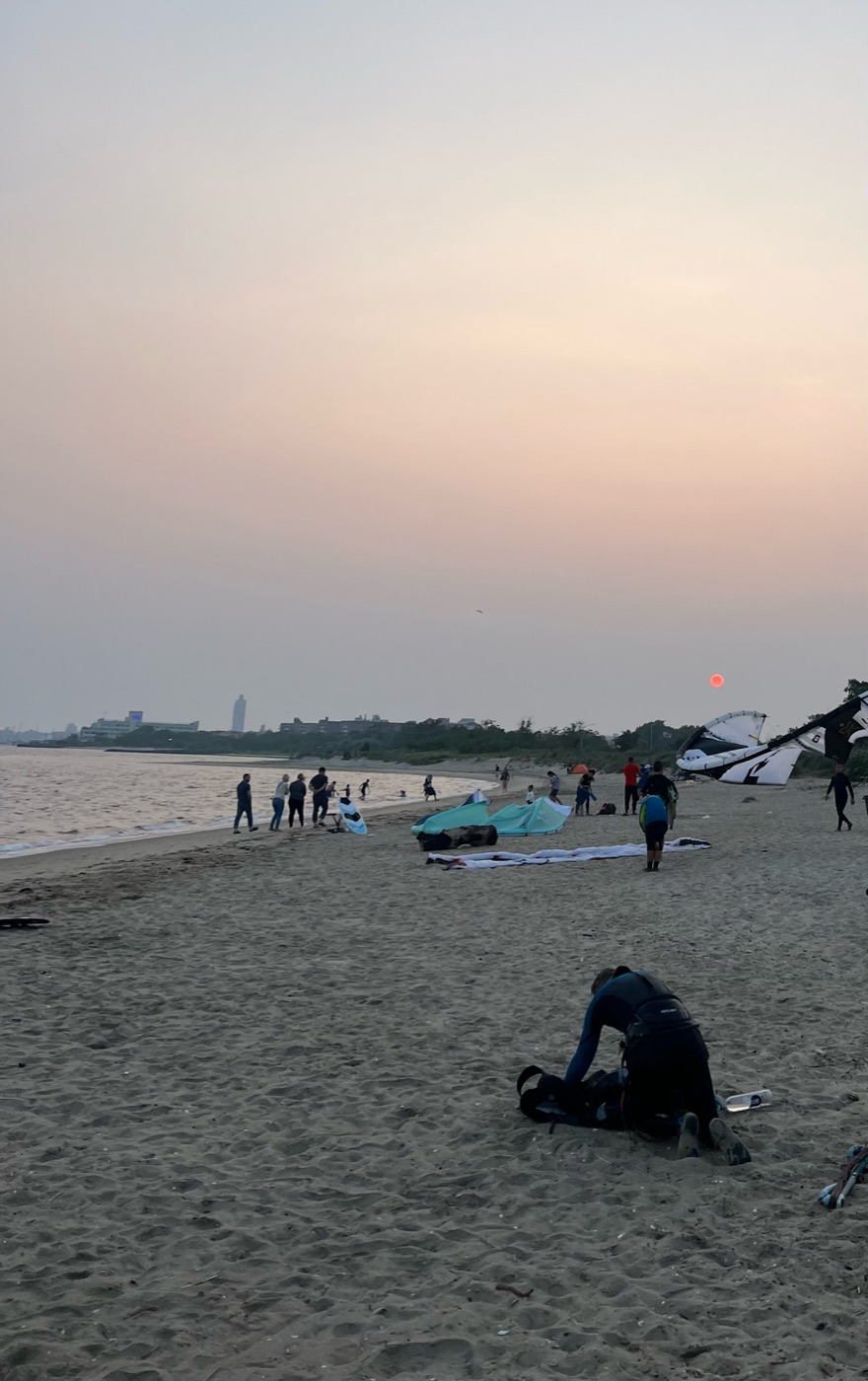 Kiteboarders pack up their gear on a dusky beach while a hazy red sun hangs low over the water.