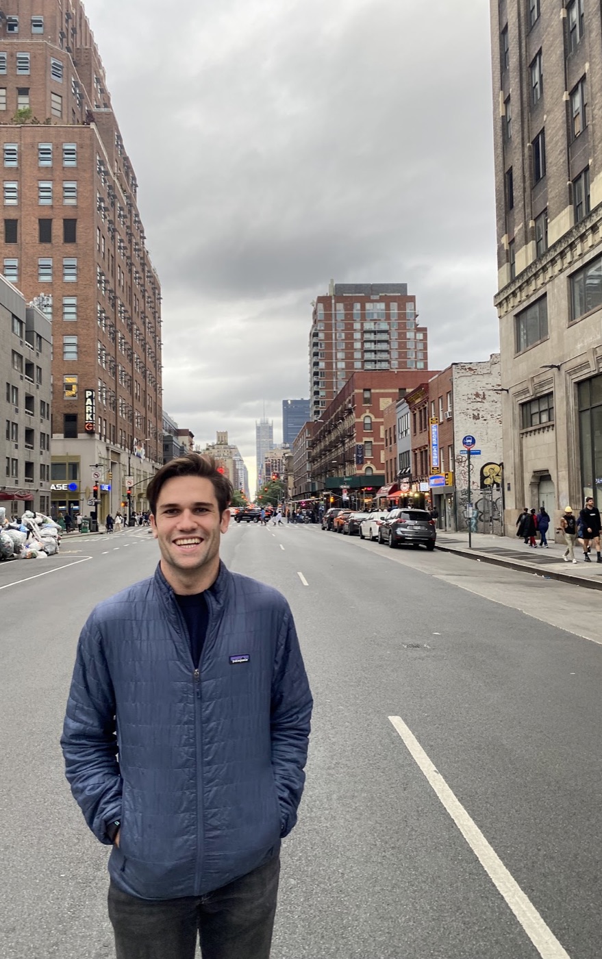 Noel stands with hands in pockets in the middle of a wide, quiet city avenue lined with brick buildings.