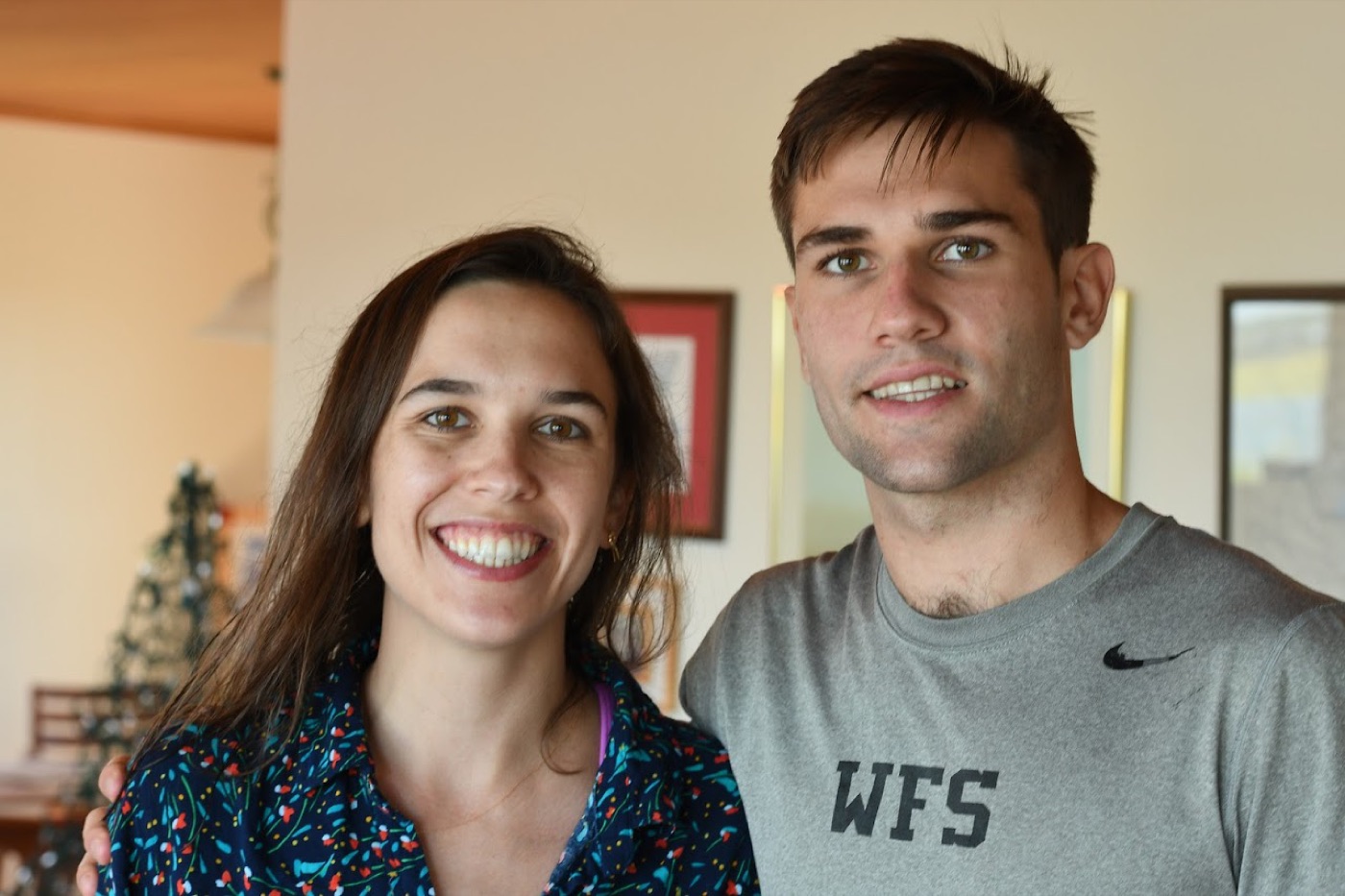 Noel in a gray WFS t-shirt stands beside a smiling woman with long brown hair in a warm-toned living room, a small tabletop Christmas tree behind them.