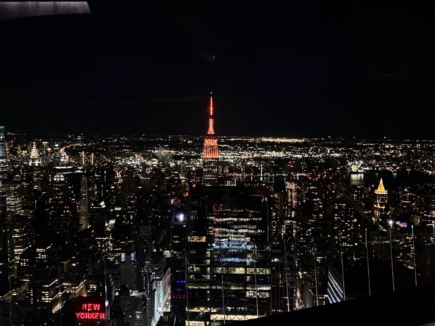 Night view over Manhattan from an observation deck, the Empire State Building's spire lit orange and the red New Yorker sign glowing far below.