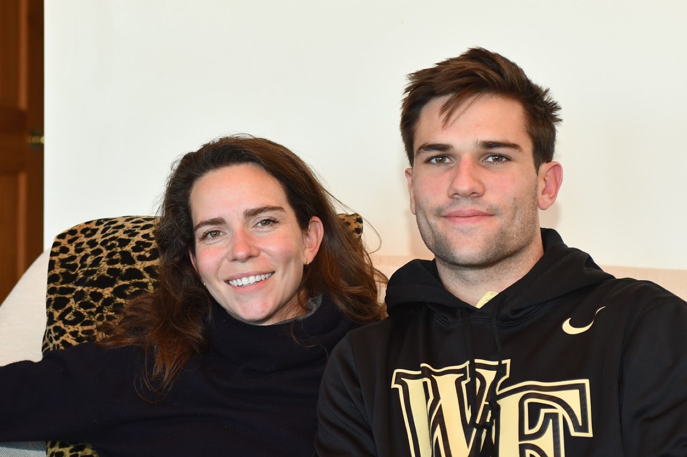 Noel in a black Wake Forest hoodie sits on a couch next to a smiling woman in a black turtleneck, a leopard-print pillow tucked behind them.