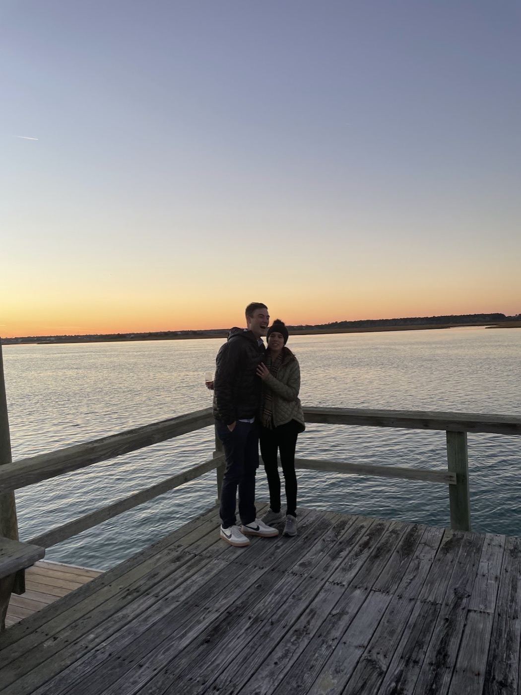 A laughing couple stands close together on a weathered wooden dock at dusk, the sun setting over calm water behind them.