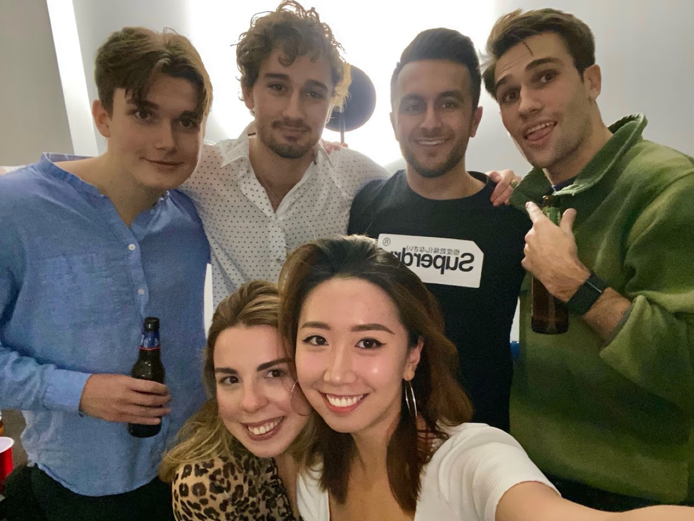 Apartment-party selfie of six friends crowded into frame, two holding beer bottles, with Noel grinning on the right in a green sweater.