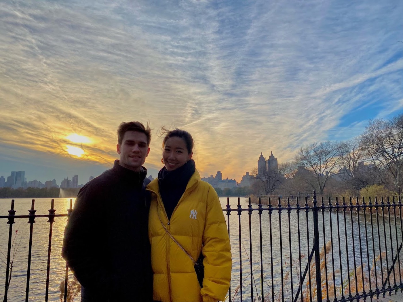 Noel and a woman in a yellow Yankees puffer jacket smile at the Central Park reservoir fence, a streaked sunset sky and the skyline behind them.