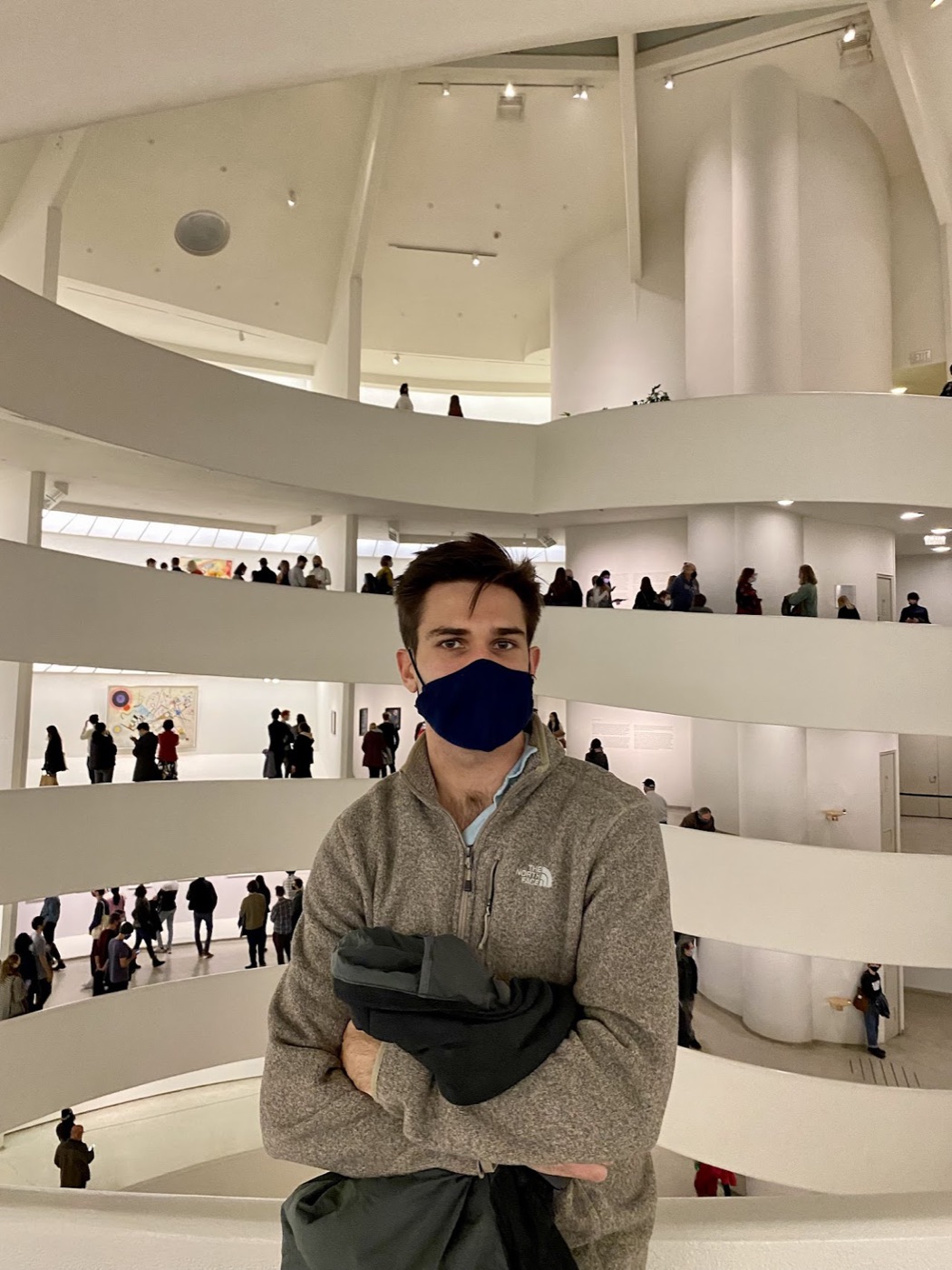 Noel, in a navy face mask and tan fleece, stands with arms crossed inside the Guggenheim's spiral rotunda while crowds browse the ramps behind him.