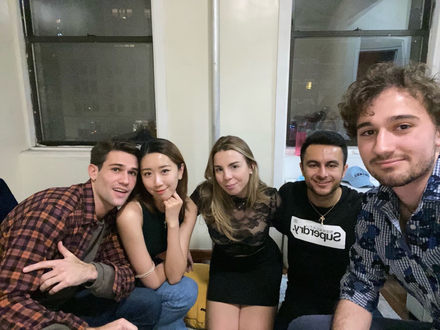 Noel in a plaid flannel flashes a hand sign in a couch selfie with four friends at a night-time apartment hangout.