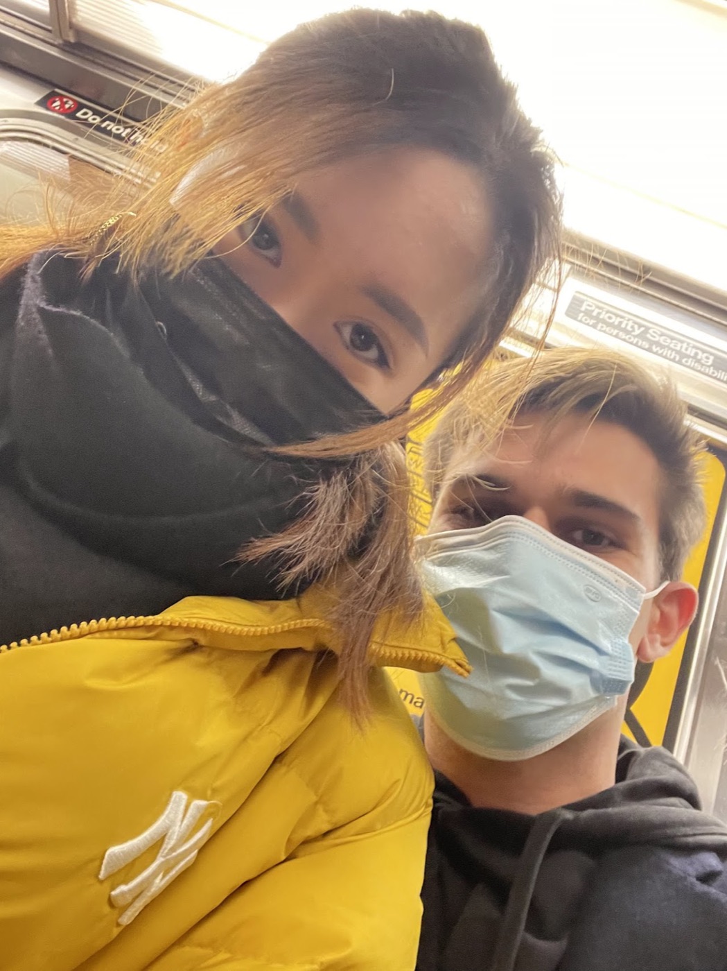 Masked subway selfie: a woman in a yellow Yankees puffer and Noel in a blue surgical mask lean into frame under a Priority Seating sign.