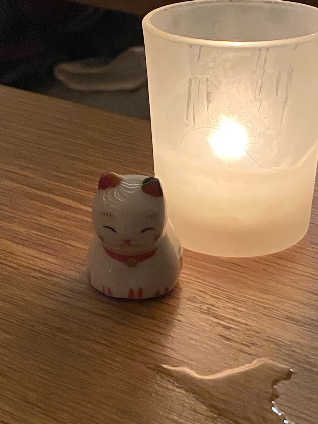 A tiny ceramic lucky-cat figurine sits on a wooden restaurant table beside a frosted glass votive glowing with candlelight.