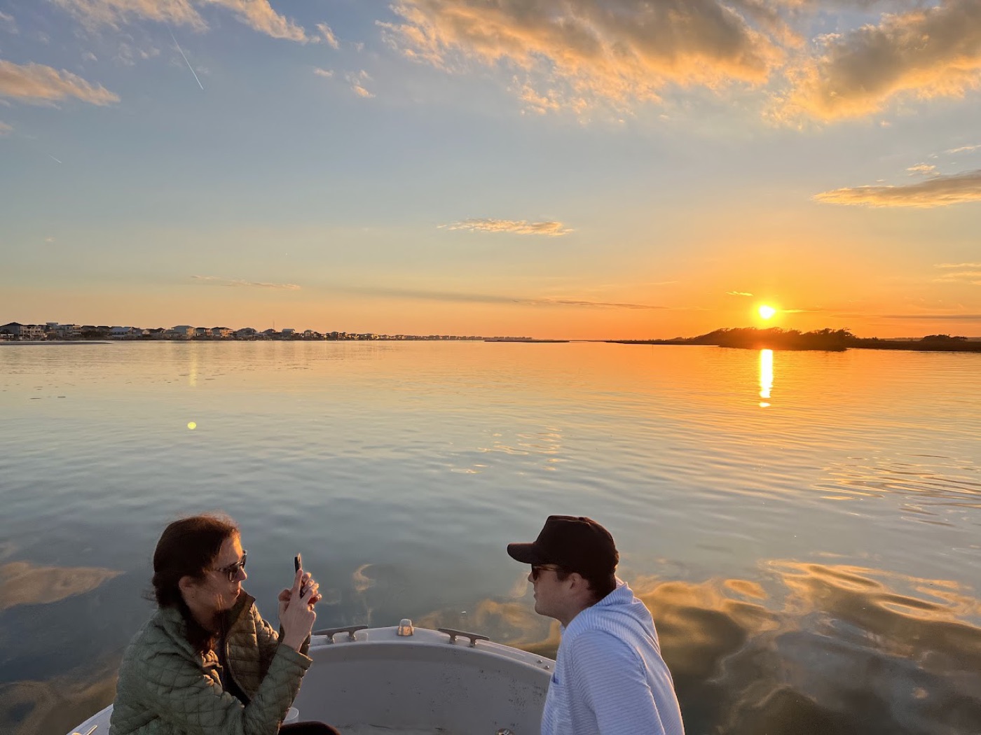 From the bow of a boat, a woman in an olive quilted jacket photographs a man in a cap and striped hoodie against a glassy orange sunset on the water.