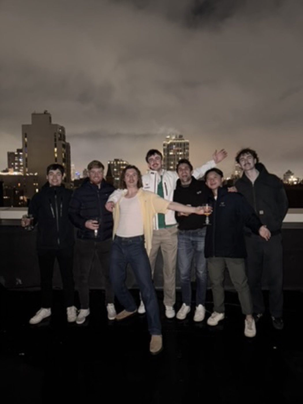 Seven friends celebrating on a rooftop against the night skyline