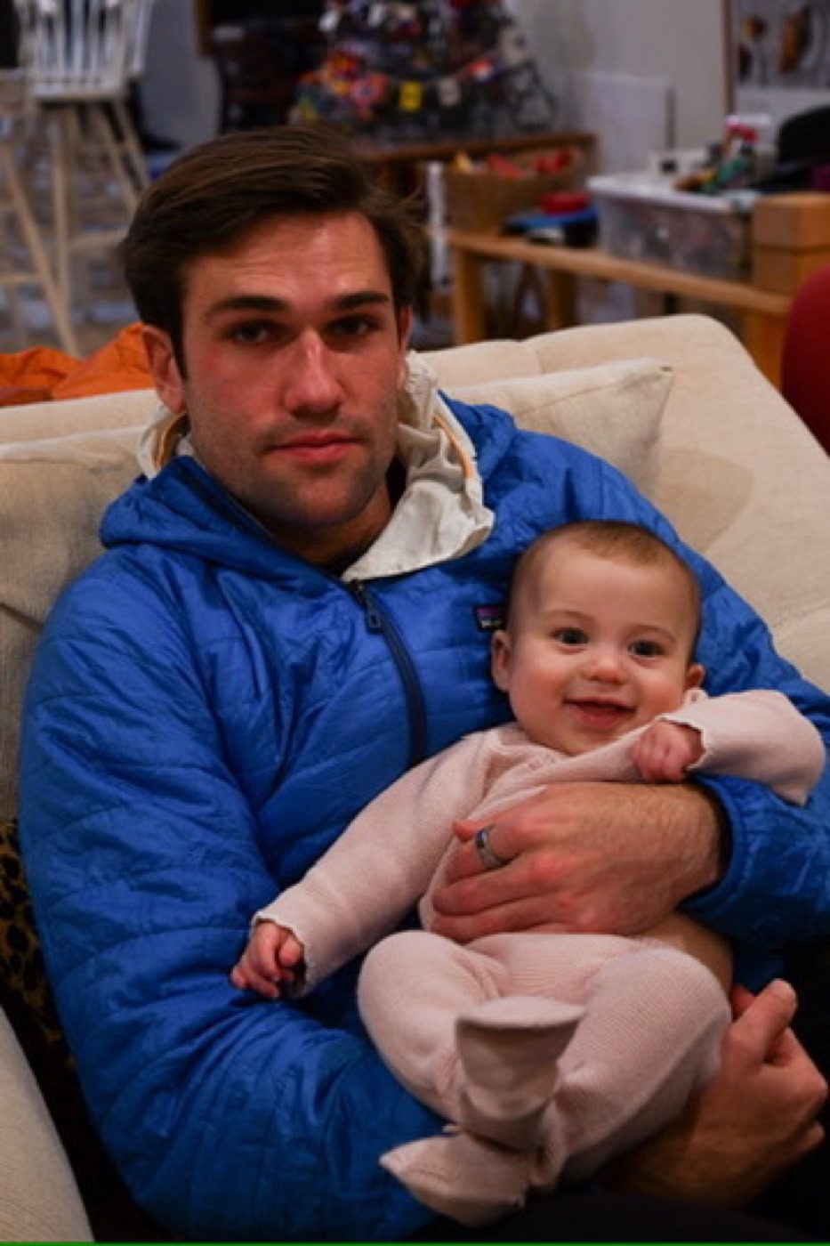 Noel cradling a beaming baby in his blue puffer indoors
