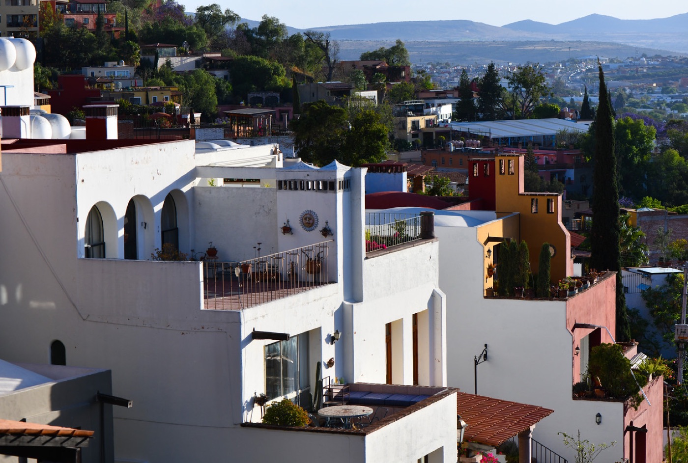 White stucco houses with rooftop terraces cascade down a hillside town toward a valley and hazy mountains in late-afternoon light.