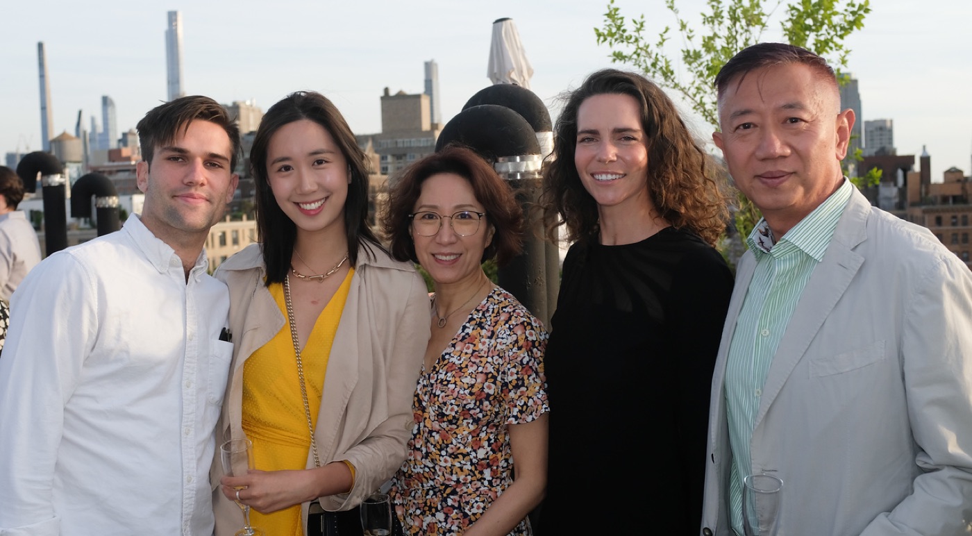 Noel and four companions share drinks at a rooftop gathering, city buildings and water towers behind them.
