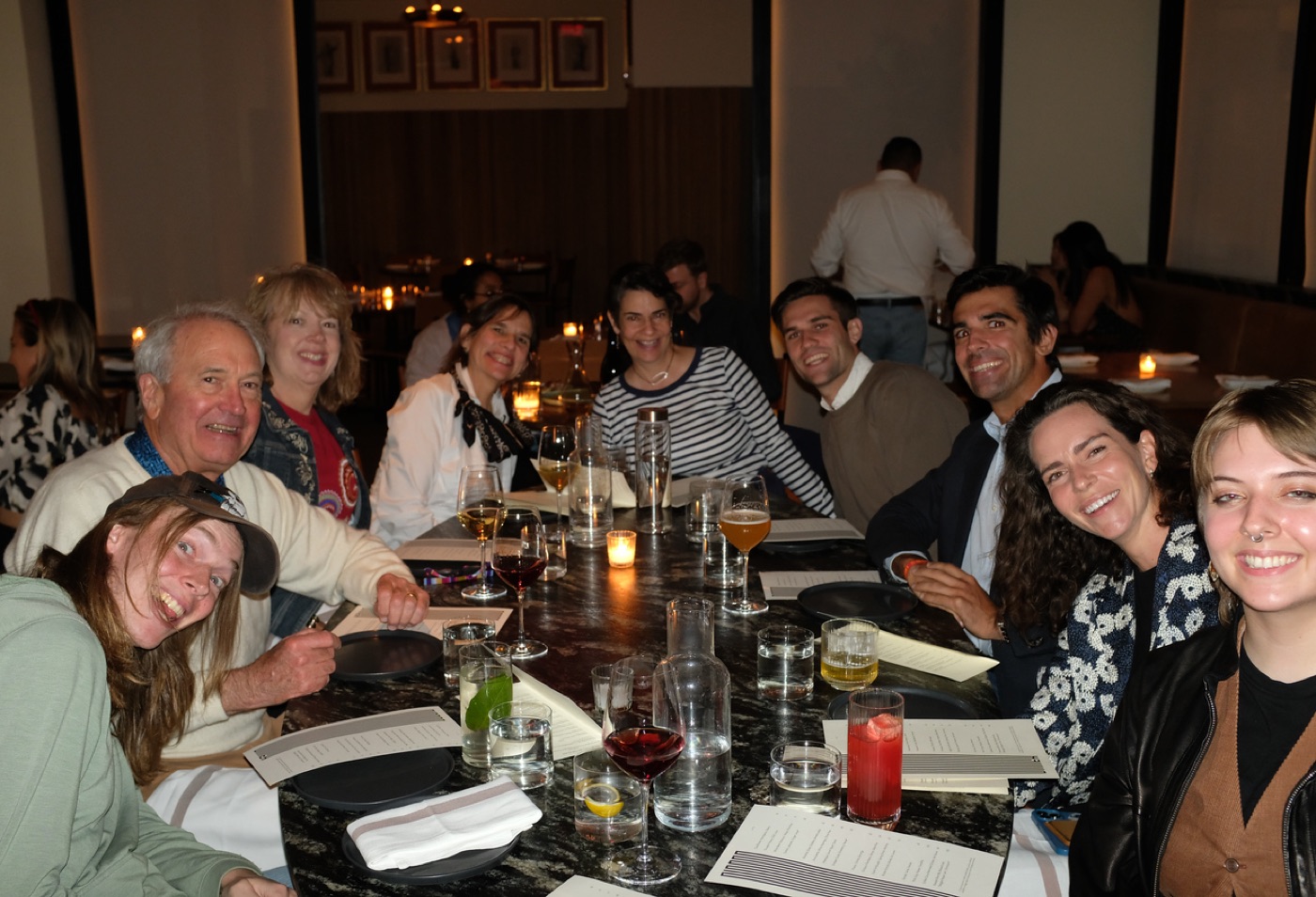 A big group of family and friends crowds around a long marble table in a dim, candlelit restaurant, smiling back at the camera.