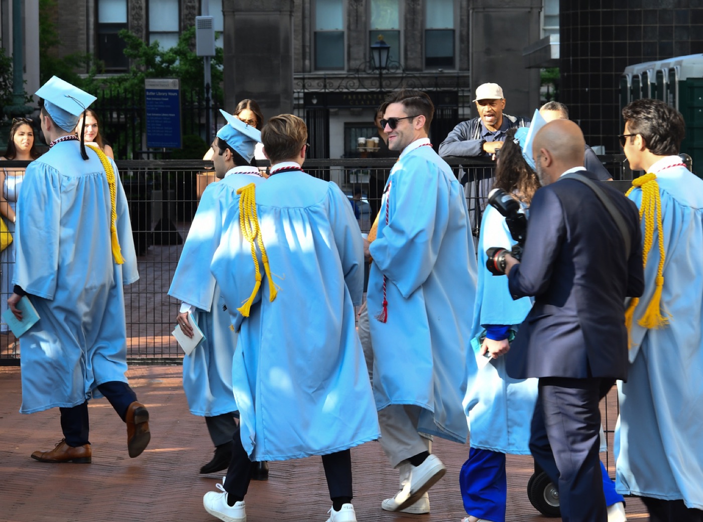 Graduates in light-blue caps and gowns with gold cords cross a brick plaza while onlookers watch from behind a fence.