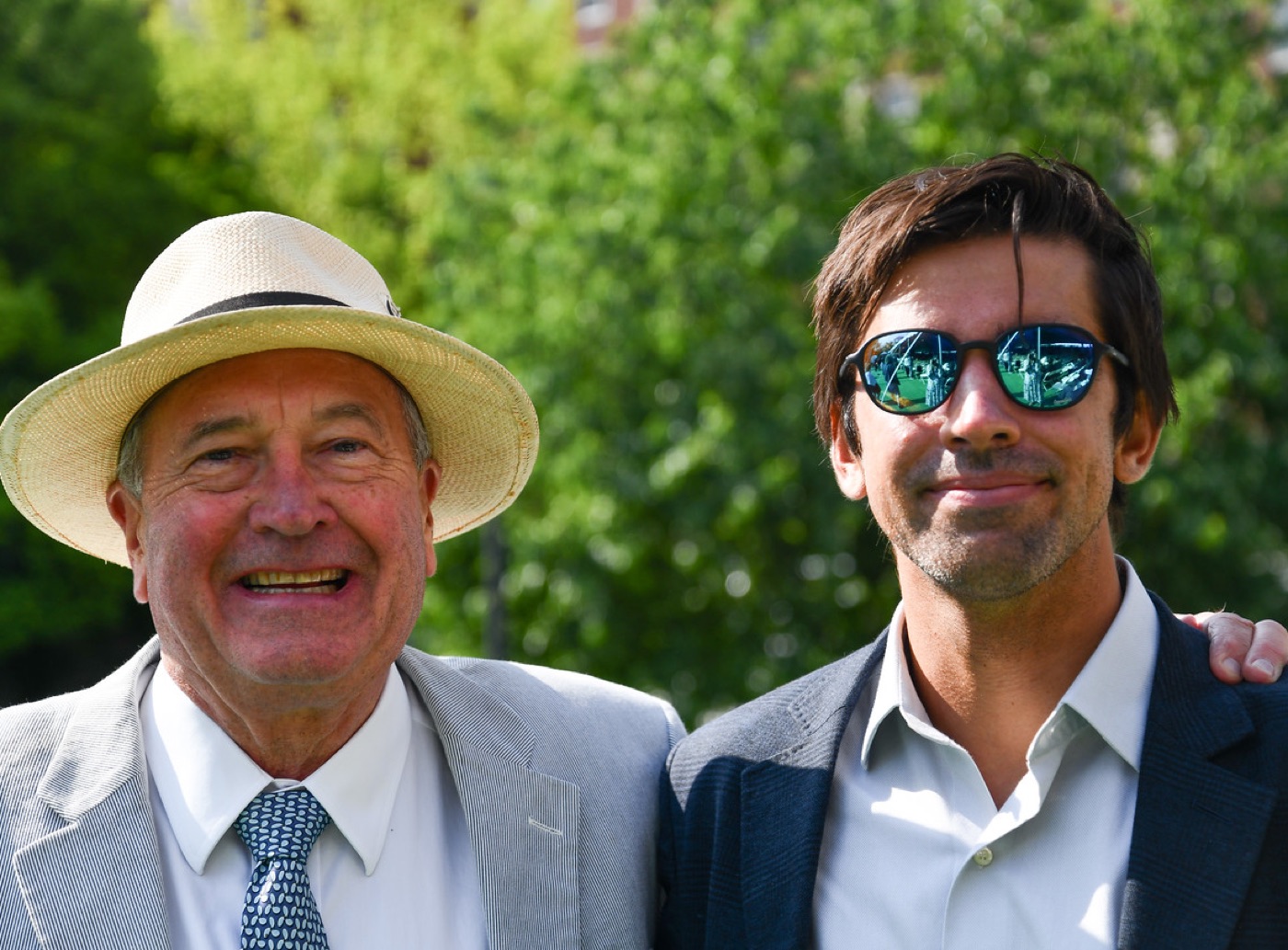 An older man in a straw hat and seersucker jacket laughs arm-in-arm with a younger man in mirrored sunglasses in front of sunlit trees.