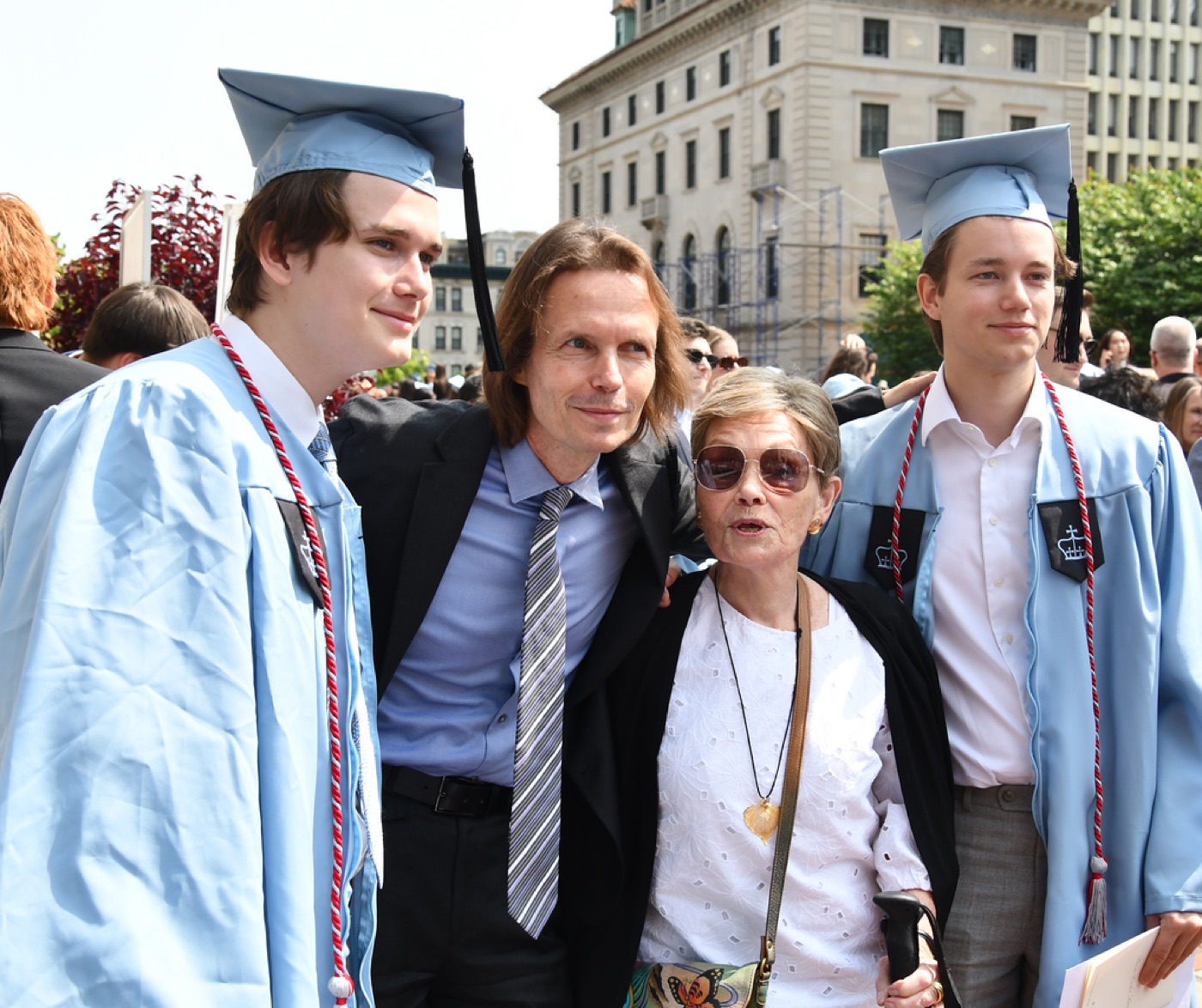 Two graduates in light-blue caps and gowns pose with a long-haired man in a suit and an older woman in a white lace top outside the ceremony.