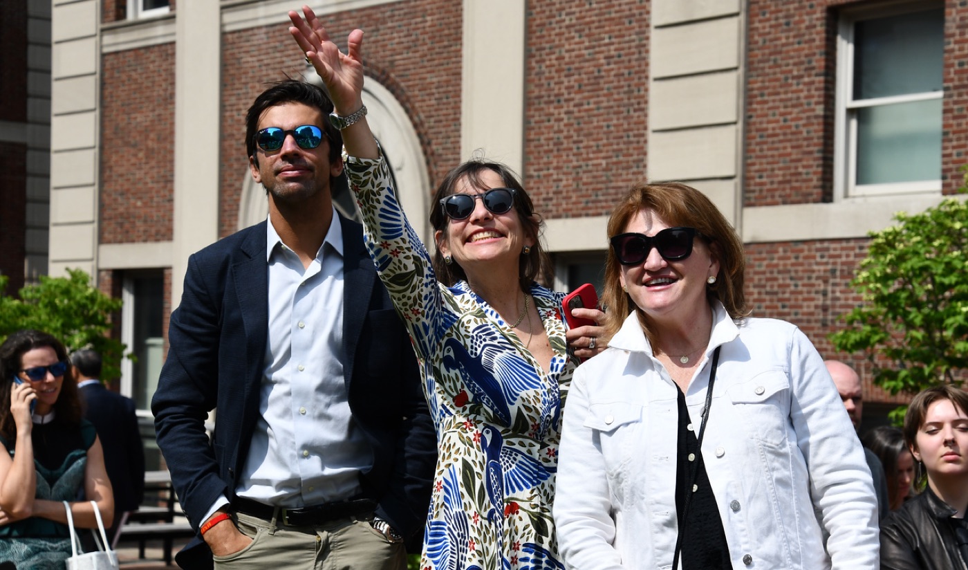A woman in a blue bird-print dress waves skyward between a man in a navy blazer and a woman in a white denim jacket outside a brick campus building.