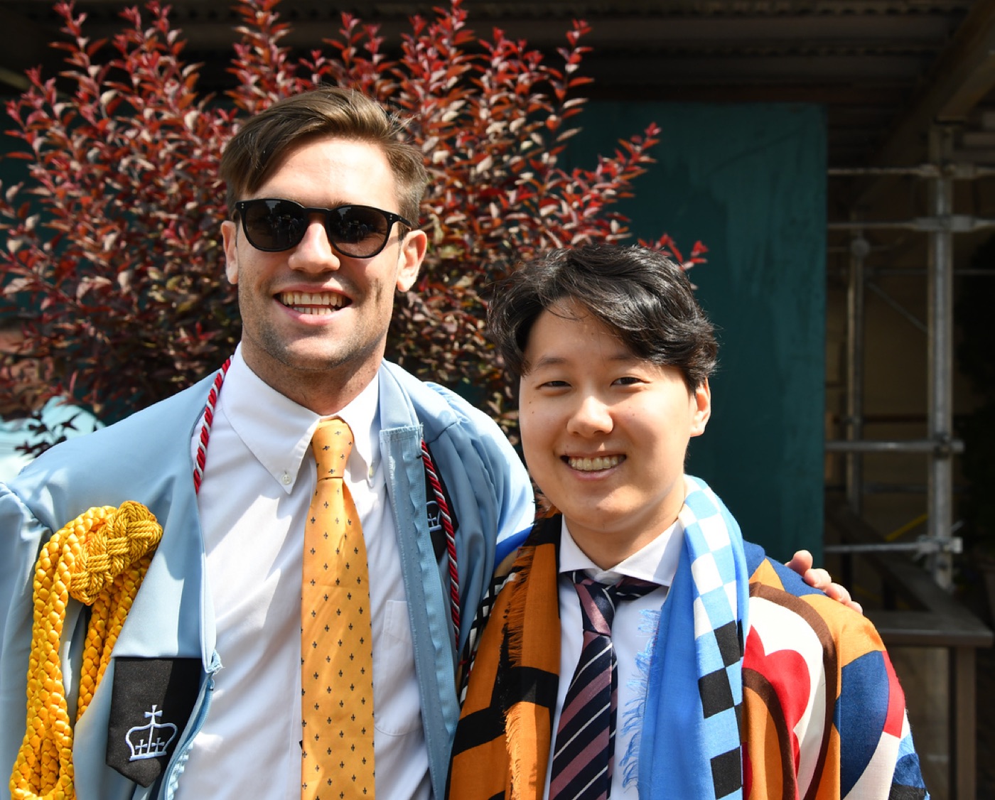 Noel, in sunglasses, a gold tie, and a light-blue Columbia graduation gown, grins beside a friend draped in a colorful scarf.