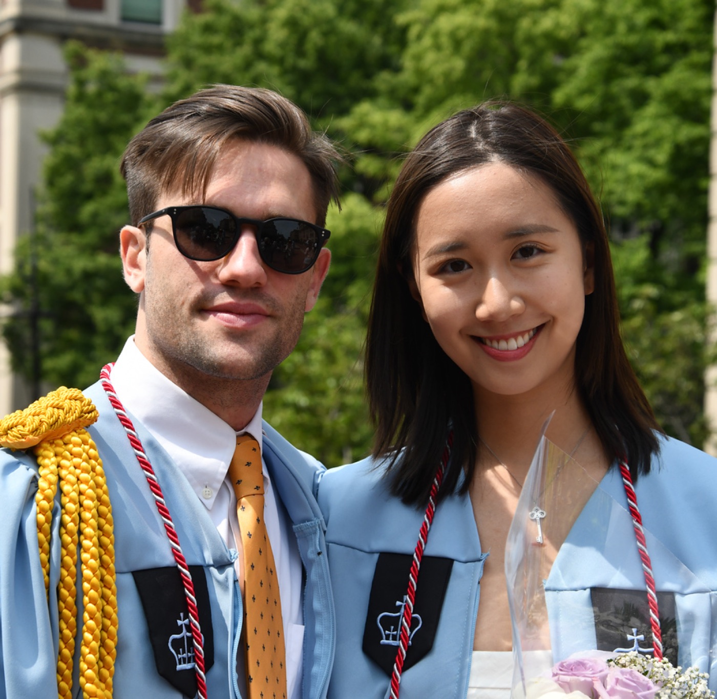 Close-up of Noel in sunglasses and a gold patterned tie beside a smiling woman, both in light-blue Columbia gowns with crimson cords.