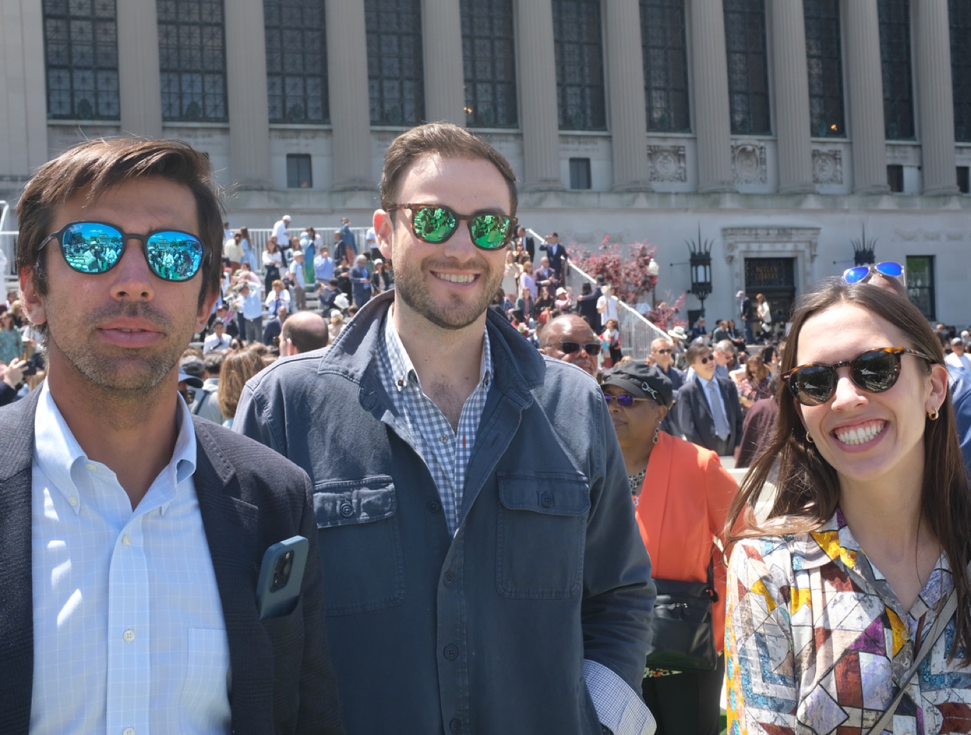 Three guests in sunglasses, two men and a smiling woman, stand in the graduation crowd in front of a columned limestone building.