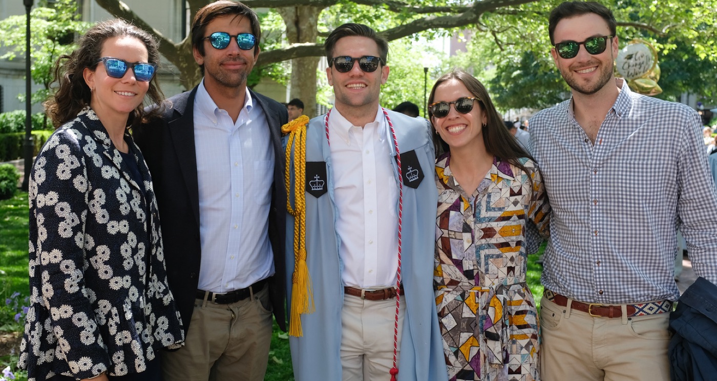 Noel, in his light-blue gown with gold and crimson cords, stands arm-in-arm with four smiling guests under a campus tree.