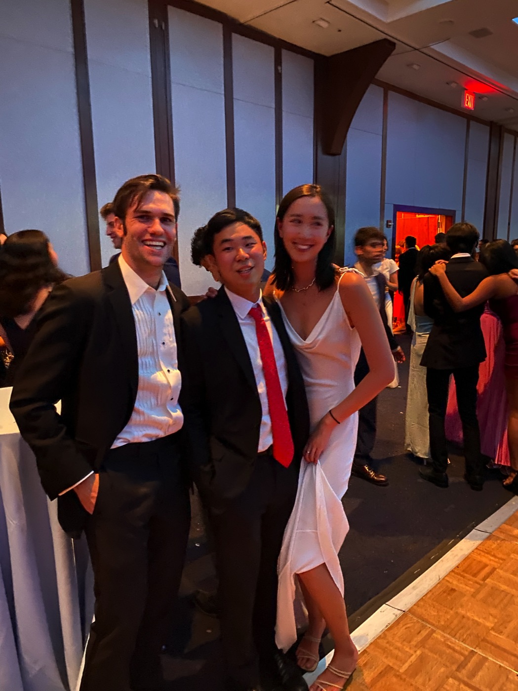 Noel in a tuxedo shirt with a friend in a red tie and a woman in a white slip dress at a formal dance, couples mingling behind them.