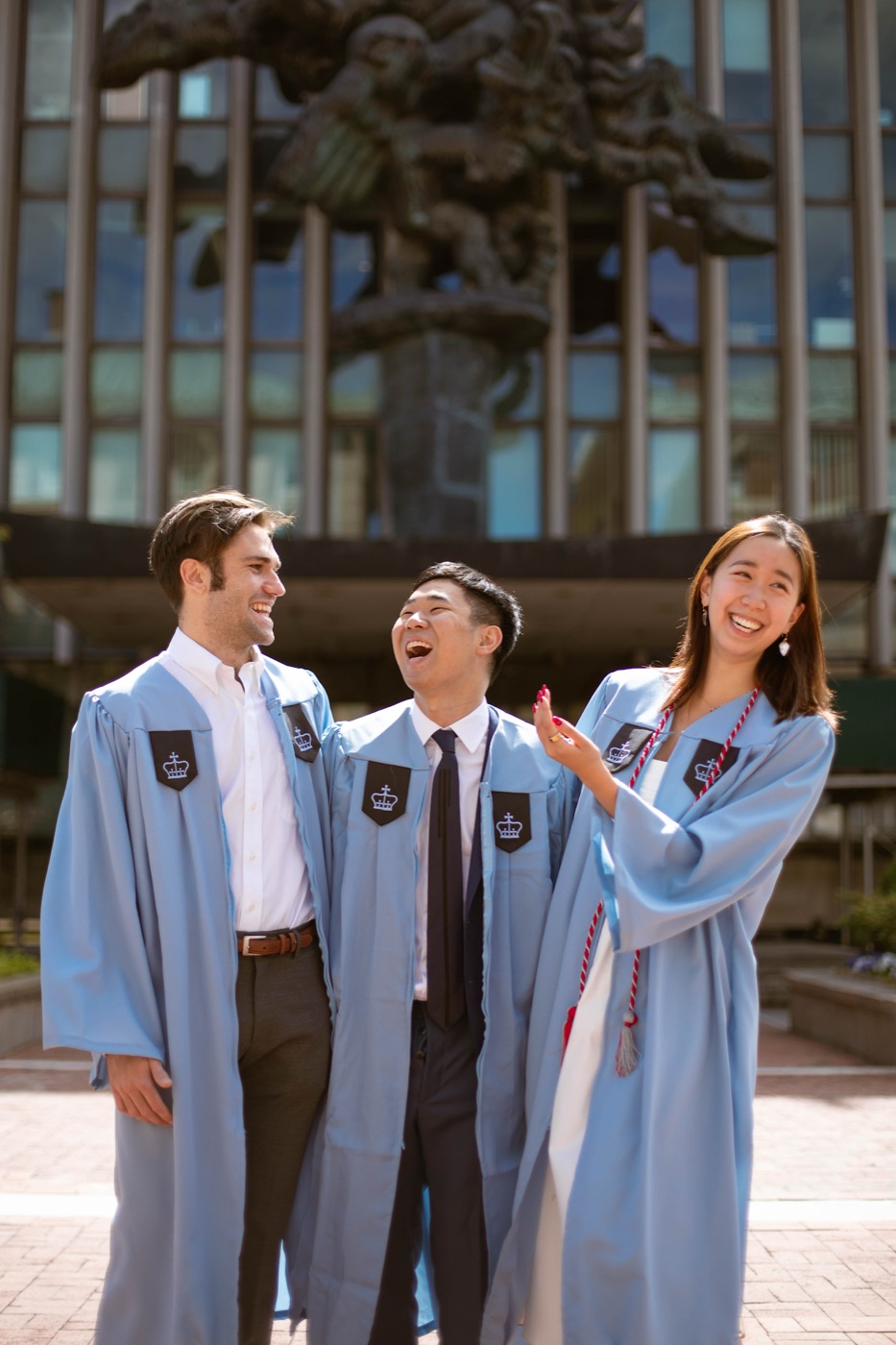 Noel and two friends in light-blue gowns crack up laughing in front of a bronze sculpture and glass facade.