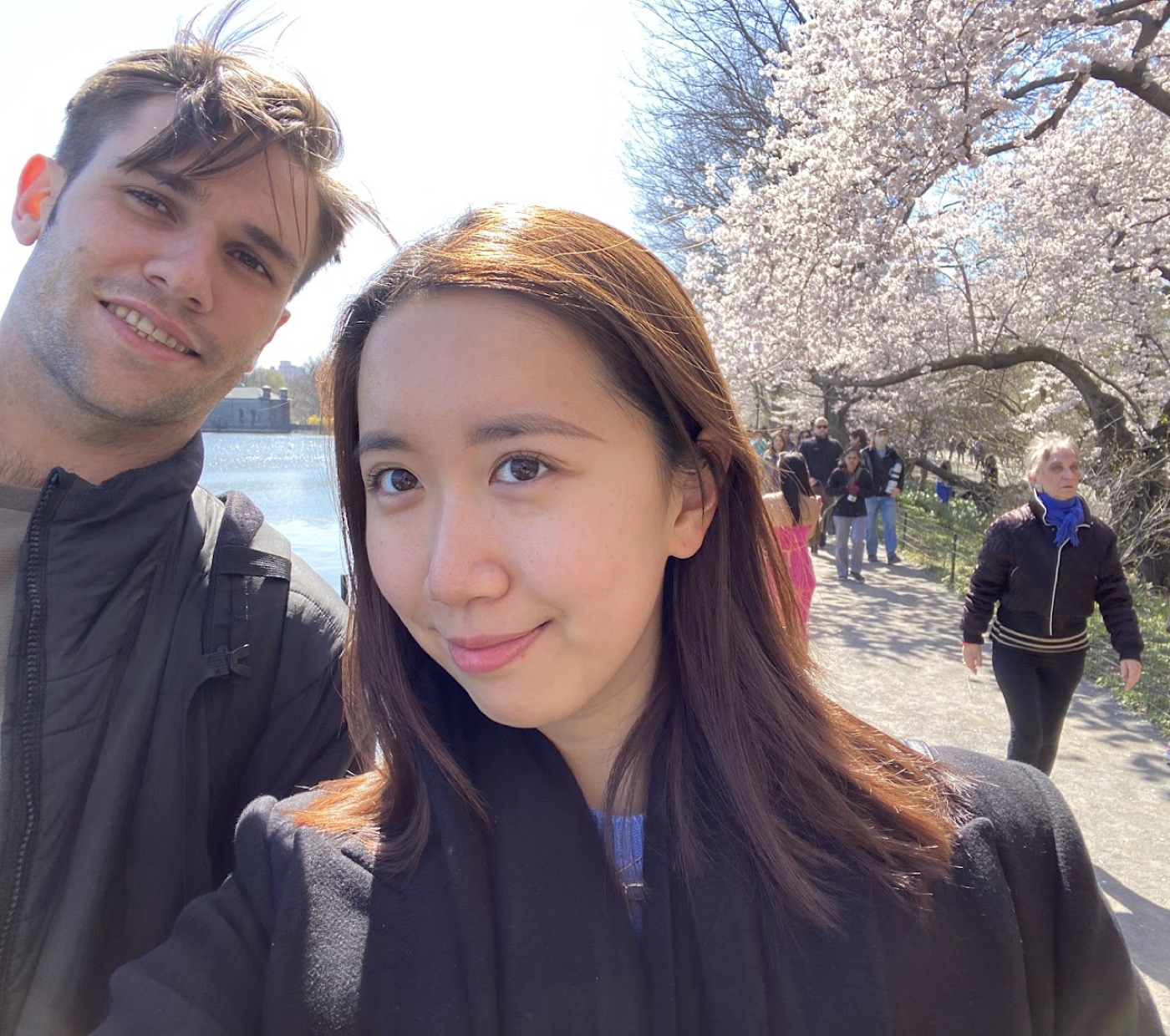 Selfie of Noel and a friend on a sunny path lined with cherry blossoms, crowds strolling under the trees behind them.