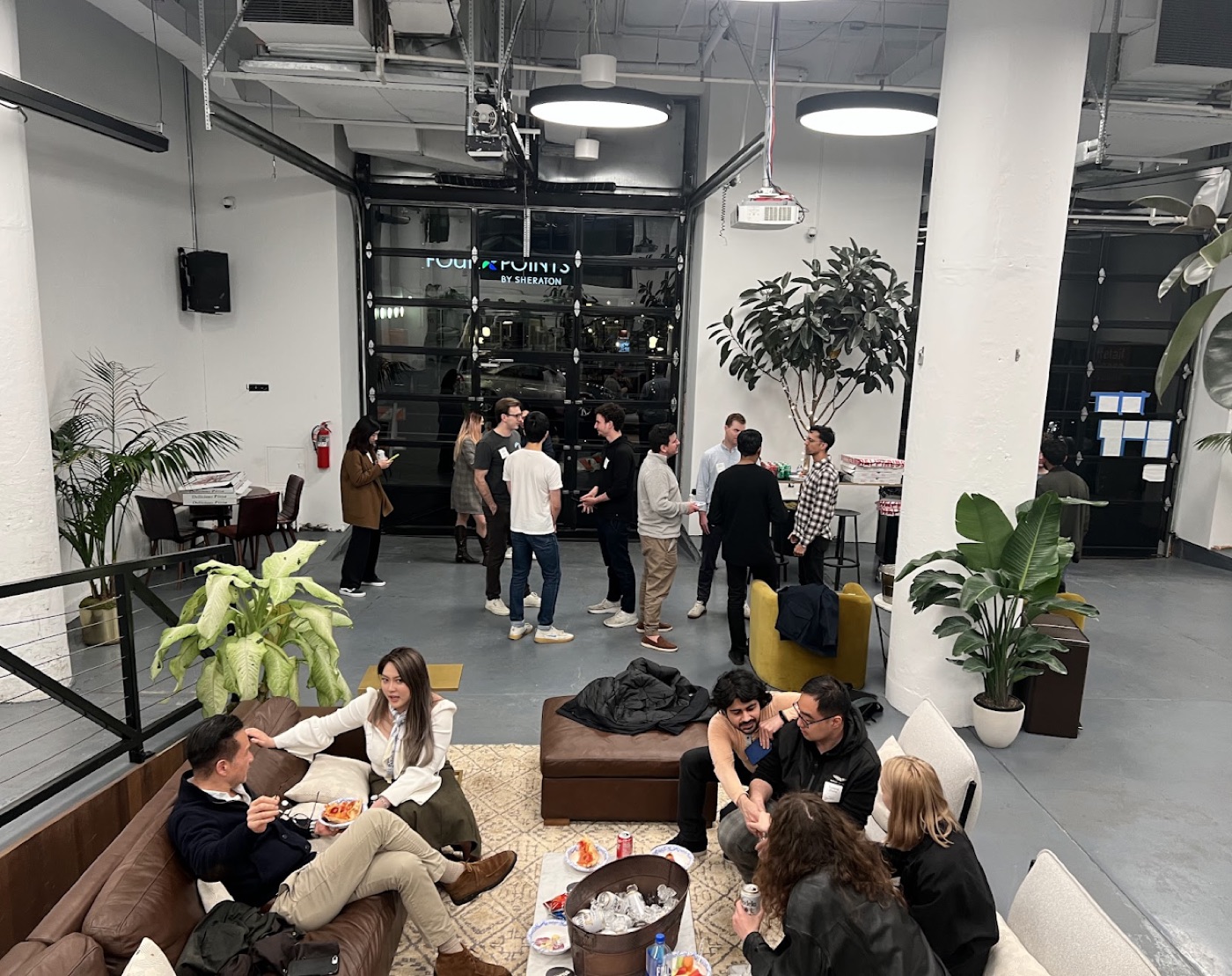 Wide view of a casual evening meetup in an industrial loft, clusters of people chatting among couches and potted plants.