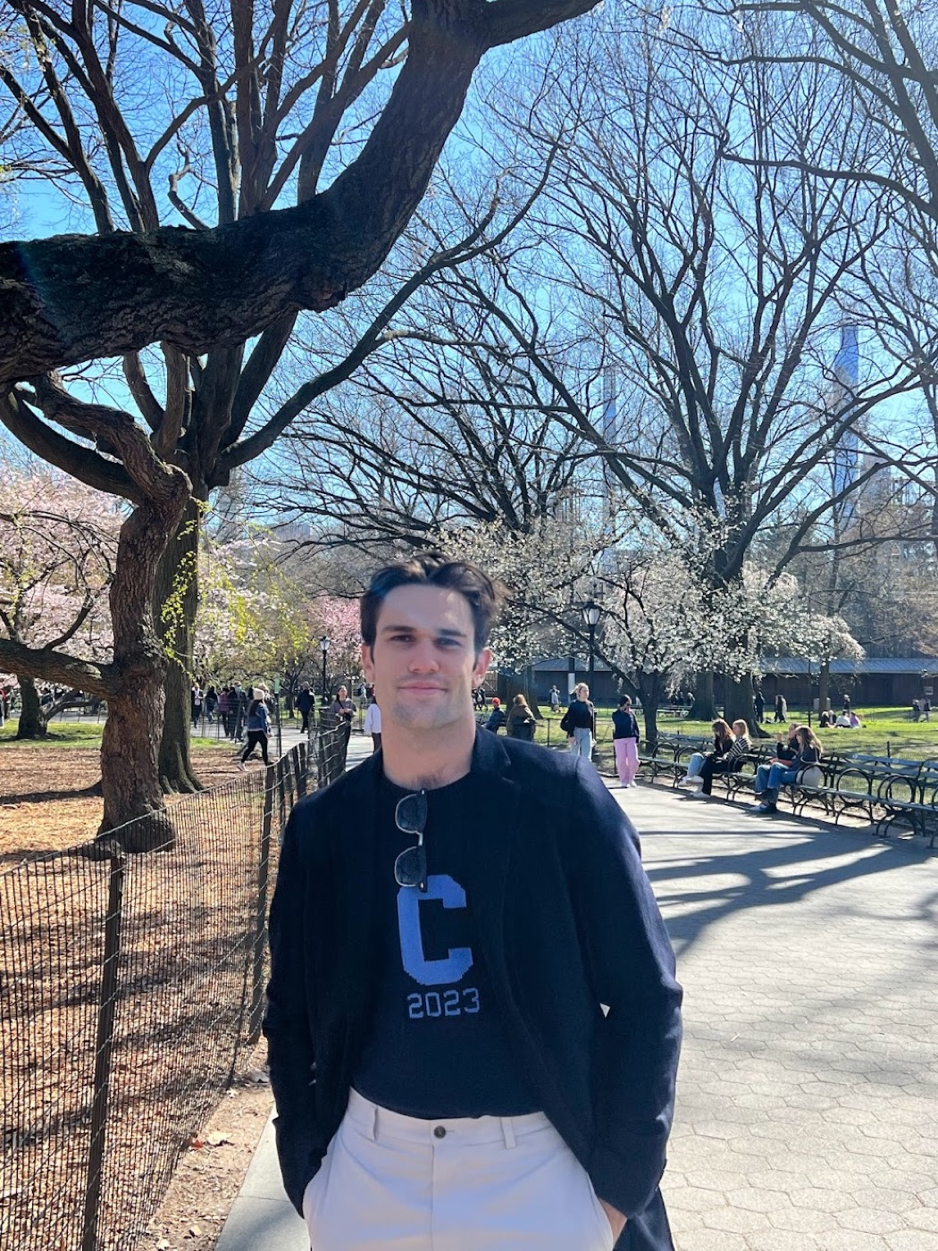 Noel stands on a park path in a navy 'C 2023' sweater and white trousers, bare trees and early blossoms behind him.
